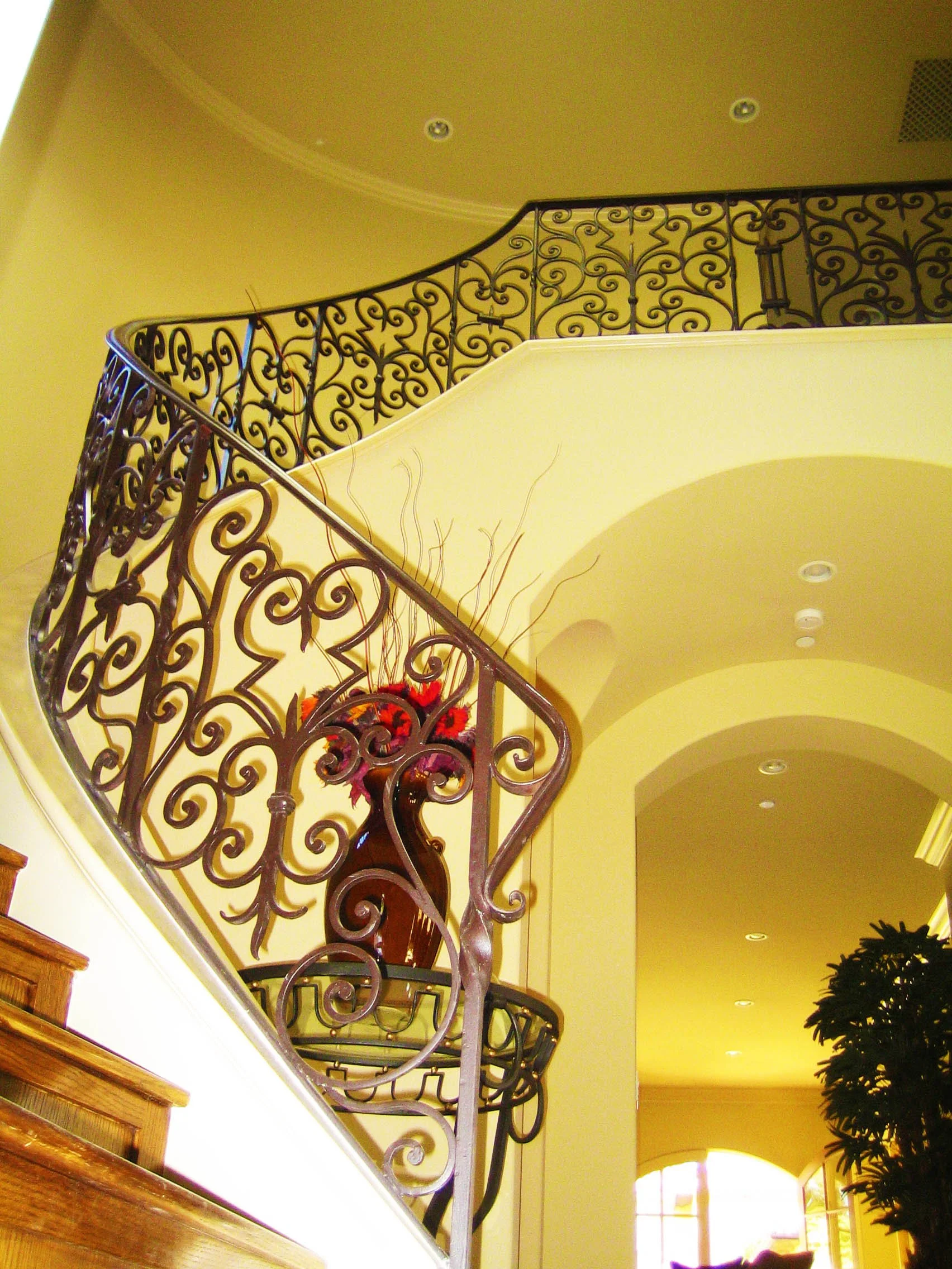Interior view of a staircase with ornate black wrought iron railing, a yellow curved wall, and a large vase with red flowers on a balcony.