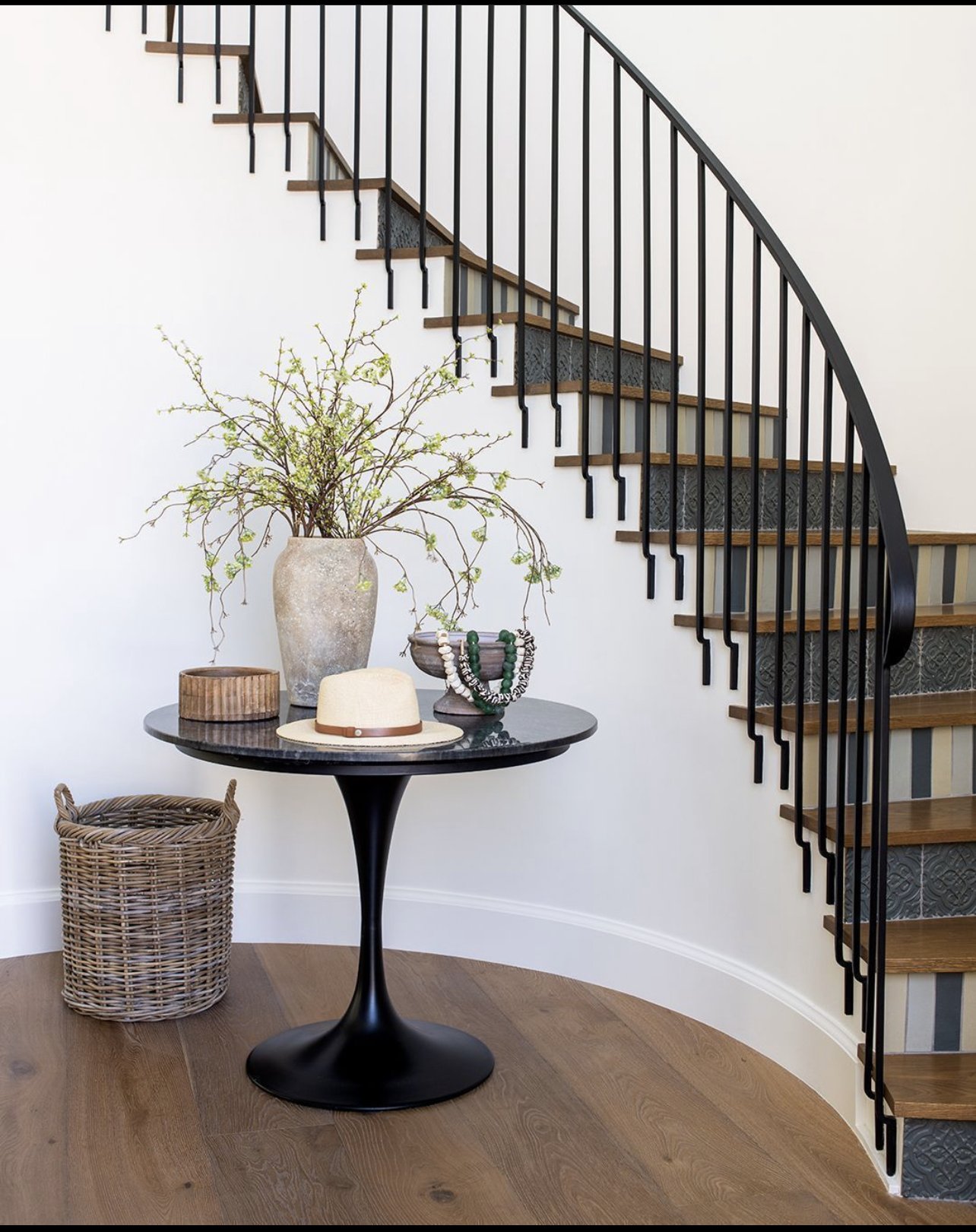 Interior decor with a black round table holding a large vase with branches, a small wooden container, a beige hat, and jewelry. Wicker basket on wooden floor next to table. Curved staircase with wooden steps and black railing in background.