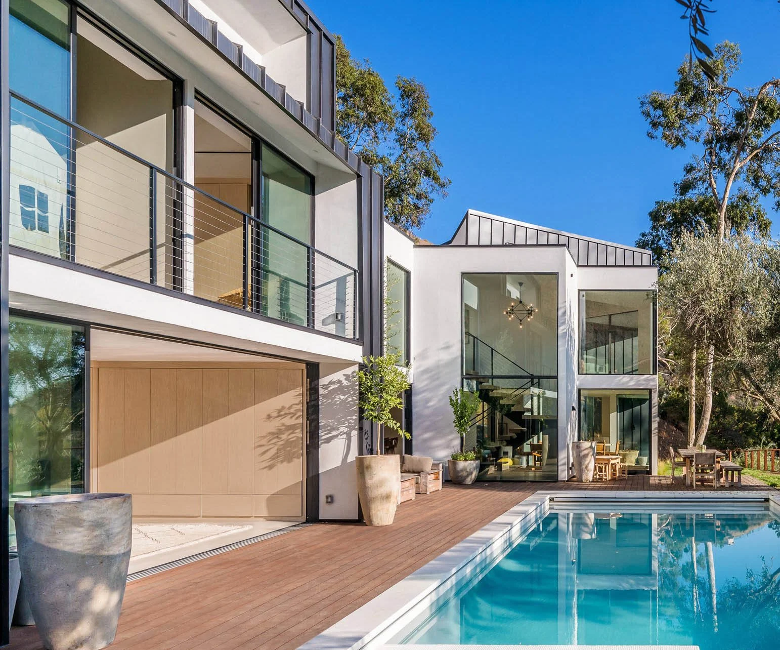 Modern white multi-story house with large glass windows and doors, a swimming pool in the foreground, wooden deck, outdoor seating, potted plants, surrounded by tall trees, under a clear blue sky.