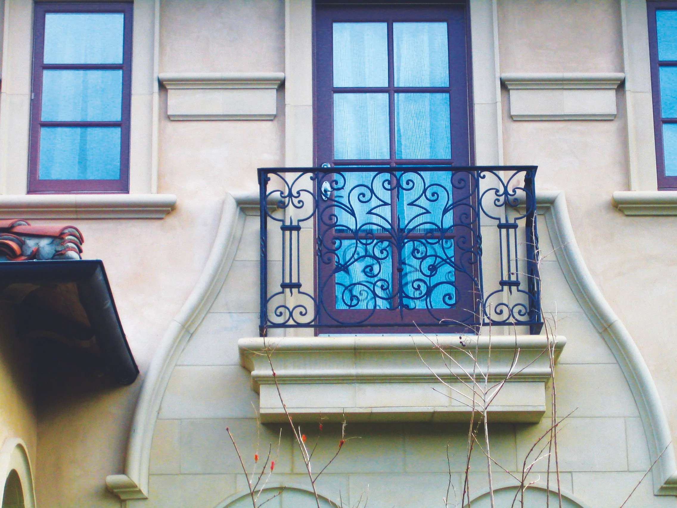 A building exterior with a small balcony featuring a decorative wrought iron railing, blue-tinted window with burgundy frame, and beige stucco wall with architectural details.