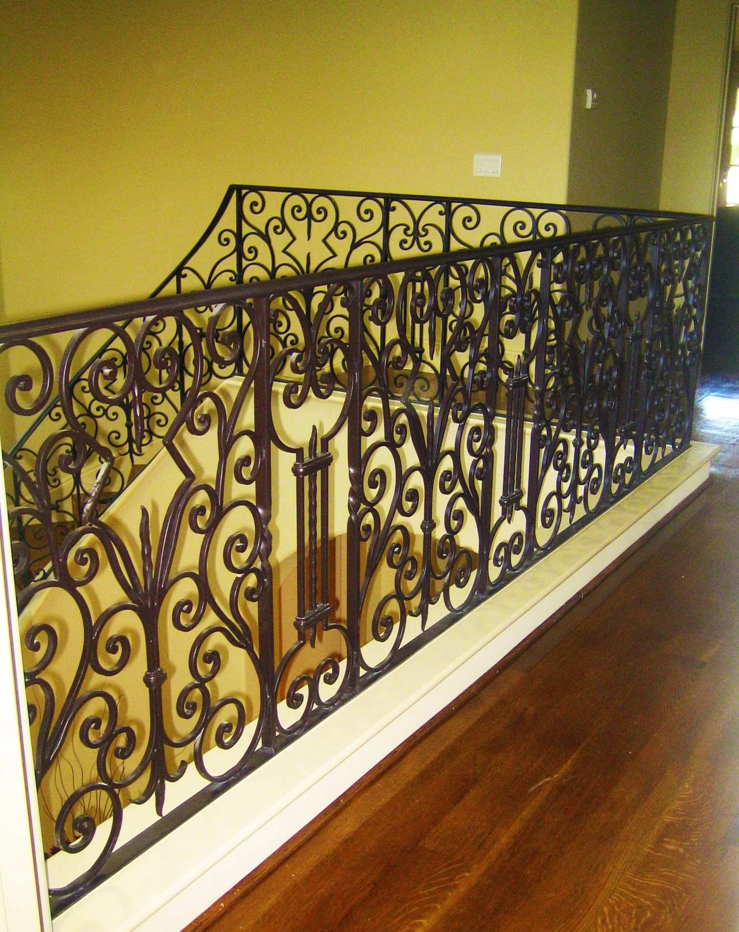 Decorative wrought iron railing along a staircase with a yellow wall behind it and a wooden floor below.