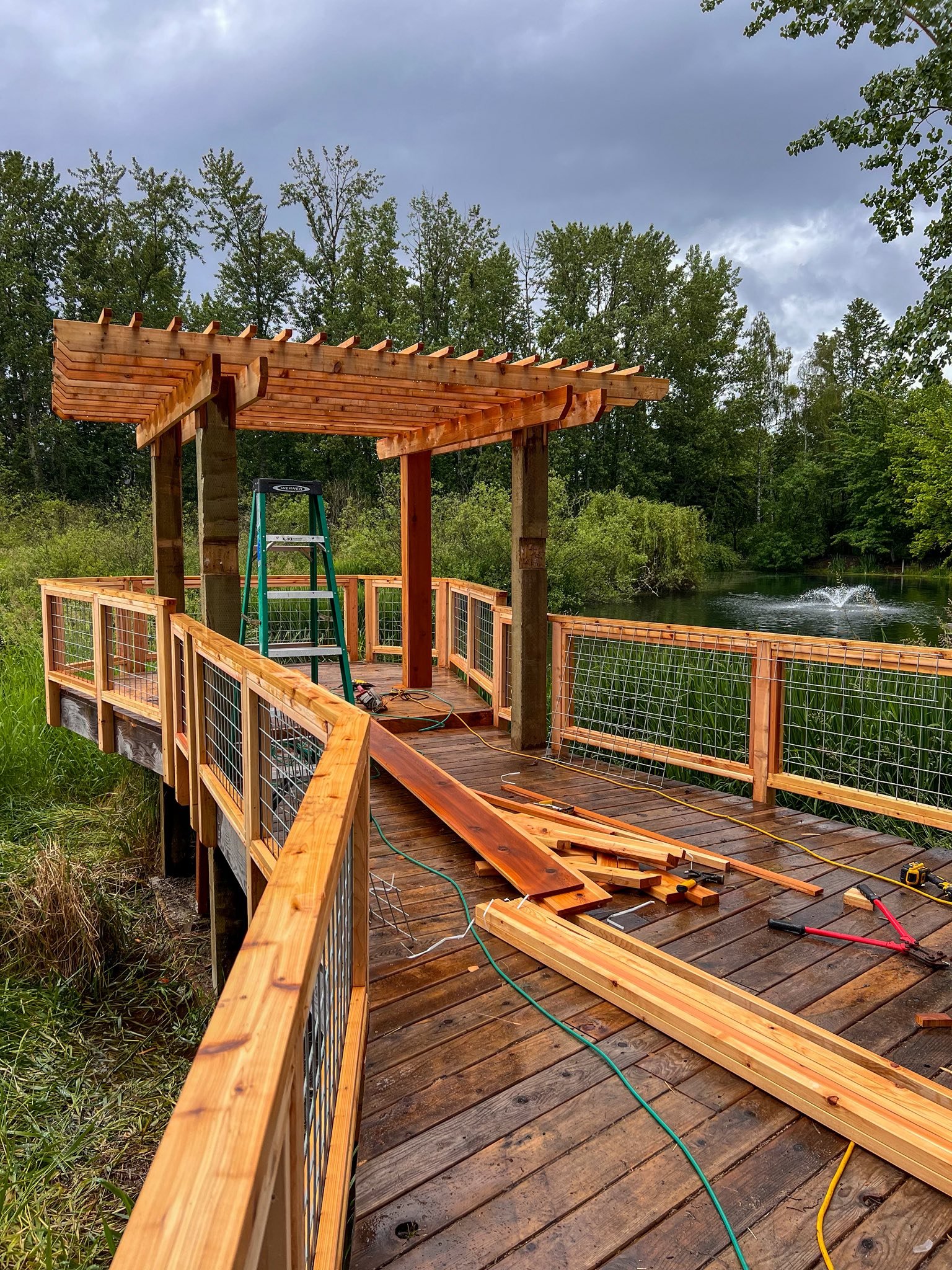 Construction of a wooden deck with a pergola near a pond, with tools and wood planks scattered on the deck.