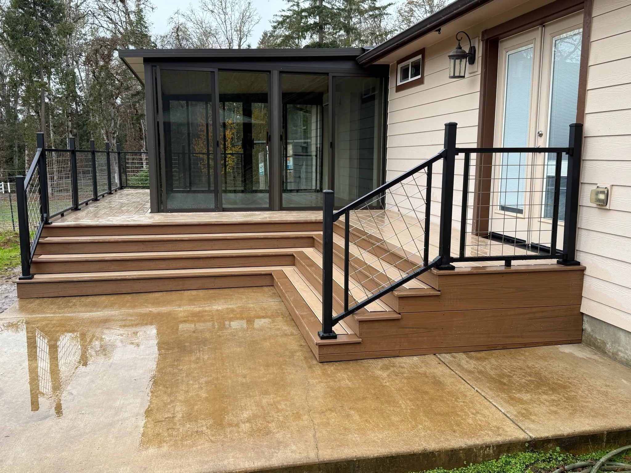 New wooden deck with stairs and black metal railings outside house with beige siding, door, and glass enclosure.