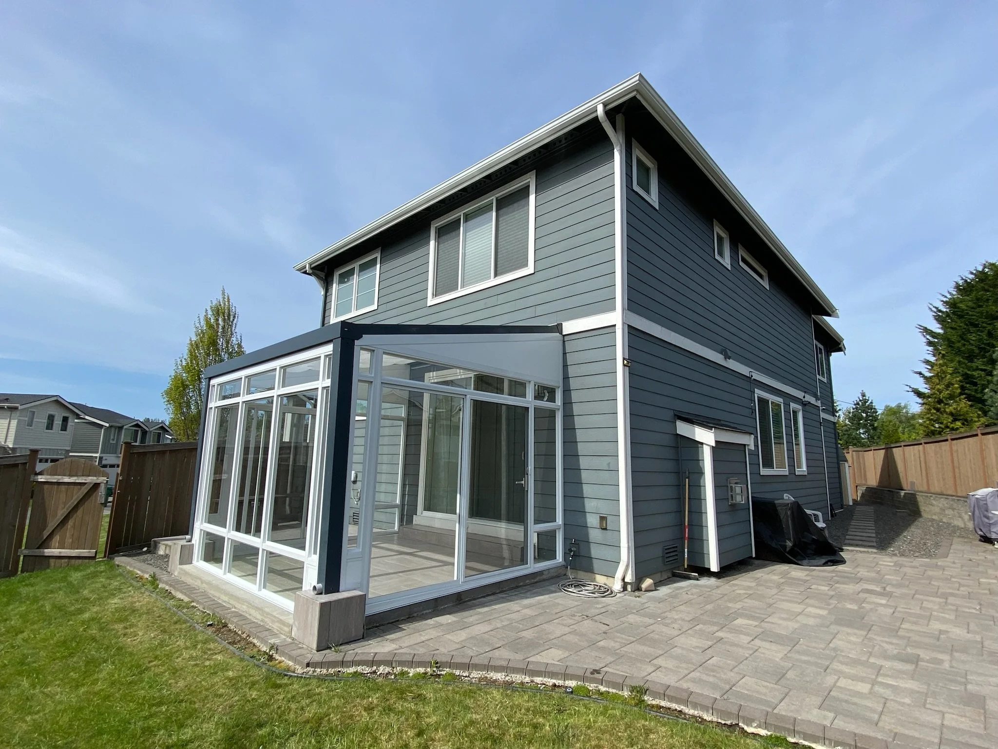 A two-story house with gray siding and white trim, featuring a glass-enclosed patio extension, in a suburban neighborhood under a blue sky.