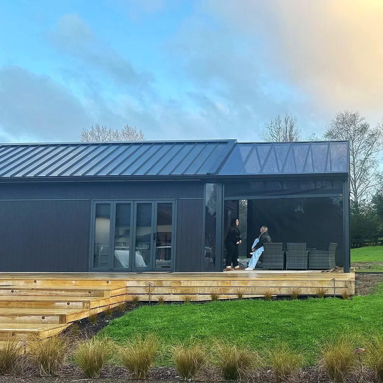 Modern black golf simulator building with a metal roof, large sliding glass doors, a wooden deck, and two people sitting and standing outside on the deck, surrounded by green grass and trees in the background under a partly cloudy sky.
