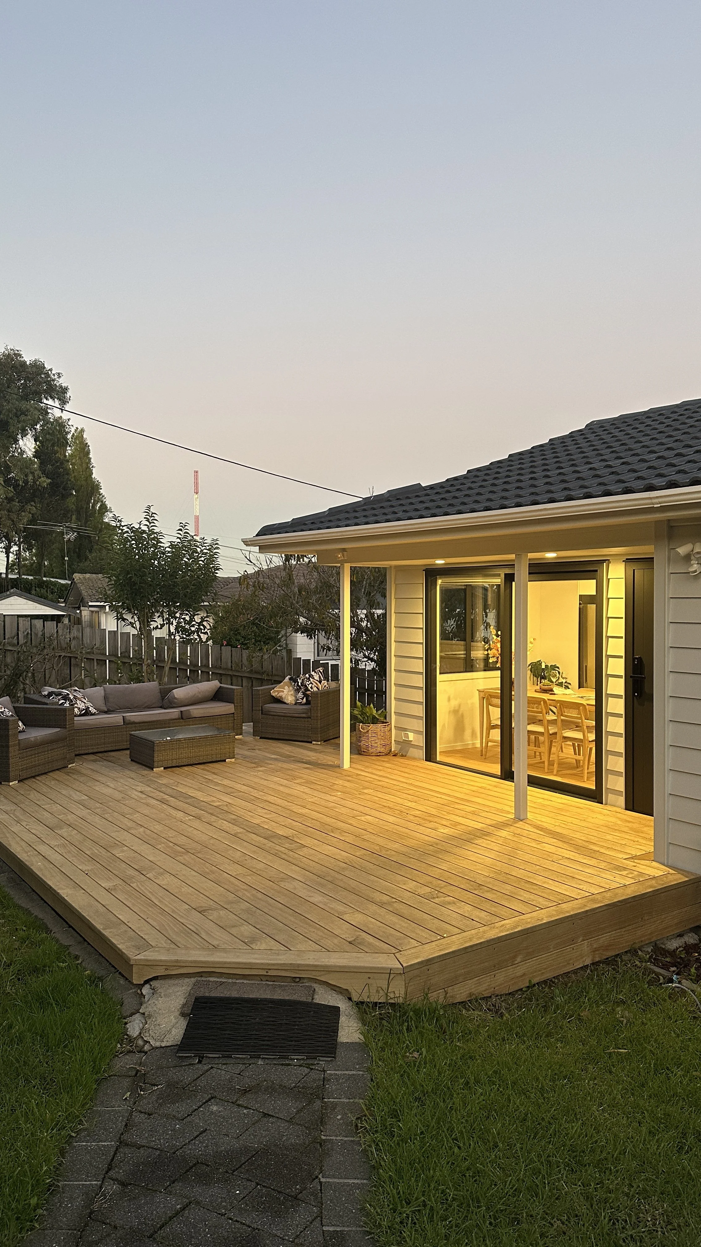 A backyard wooden deck with outdoor furniture, and a family house with large glass doors leading to a dining area, illuminated by interior lighting in the evening.