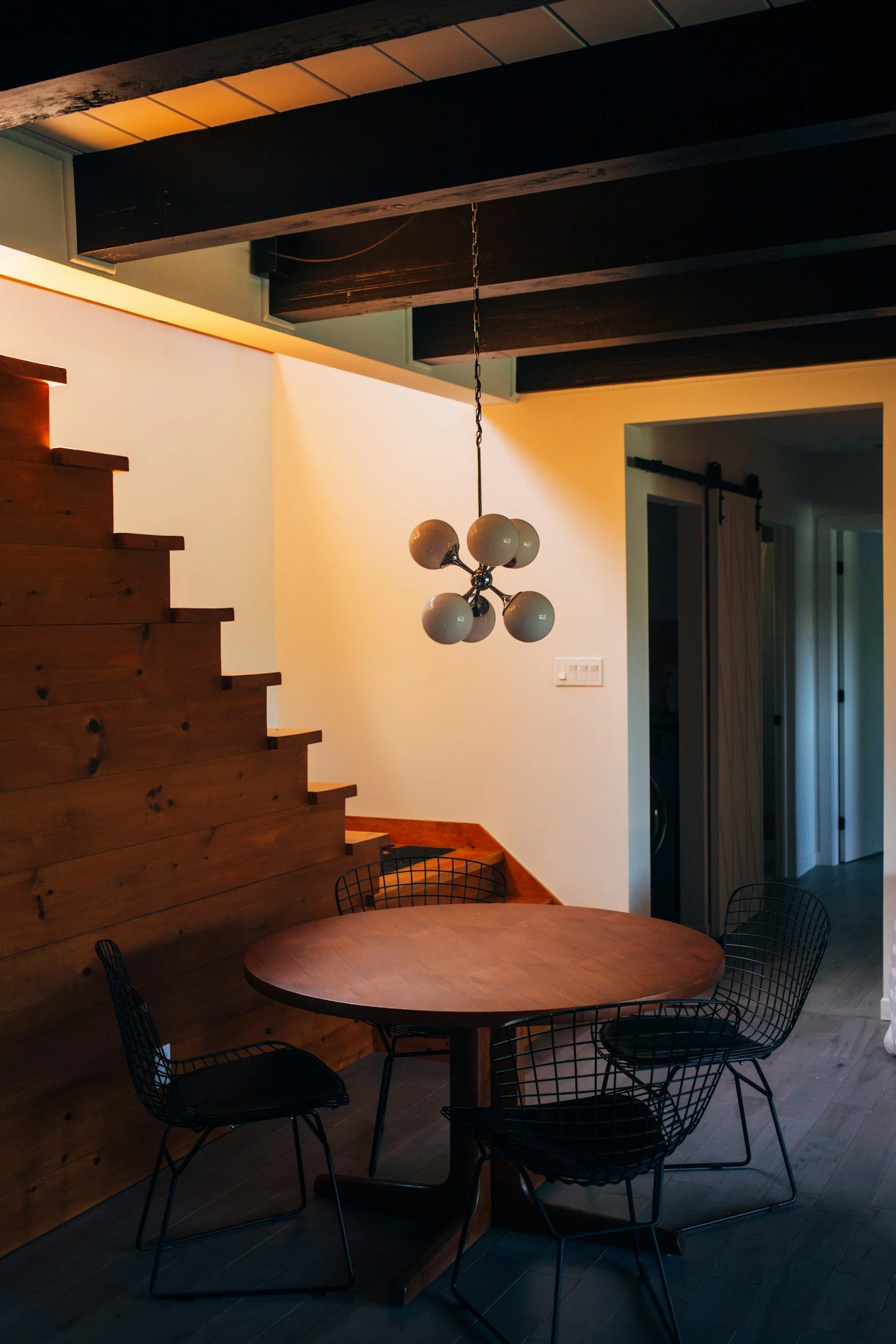 Interior of a house with wooden staircase, round dining table, black wire chairs, modern chandelier, wooden ceiling beams, and hallway with sliding barn door.