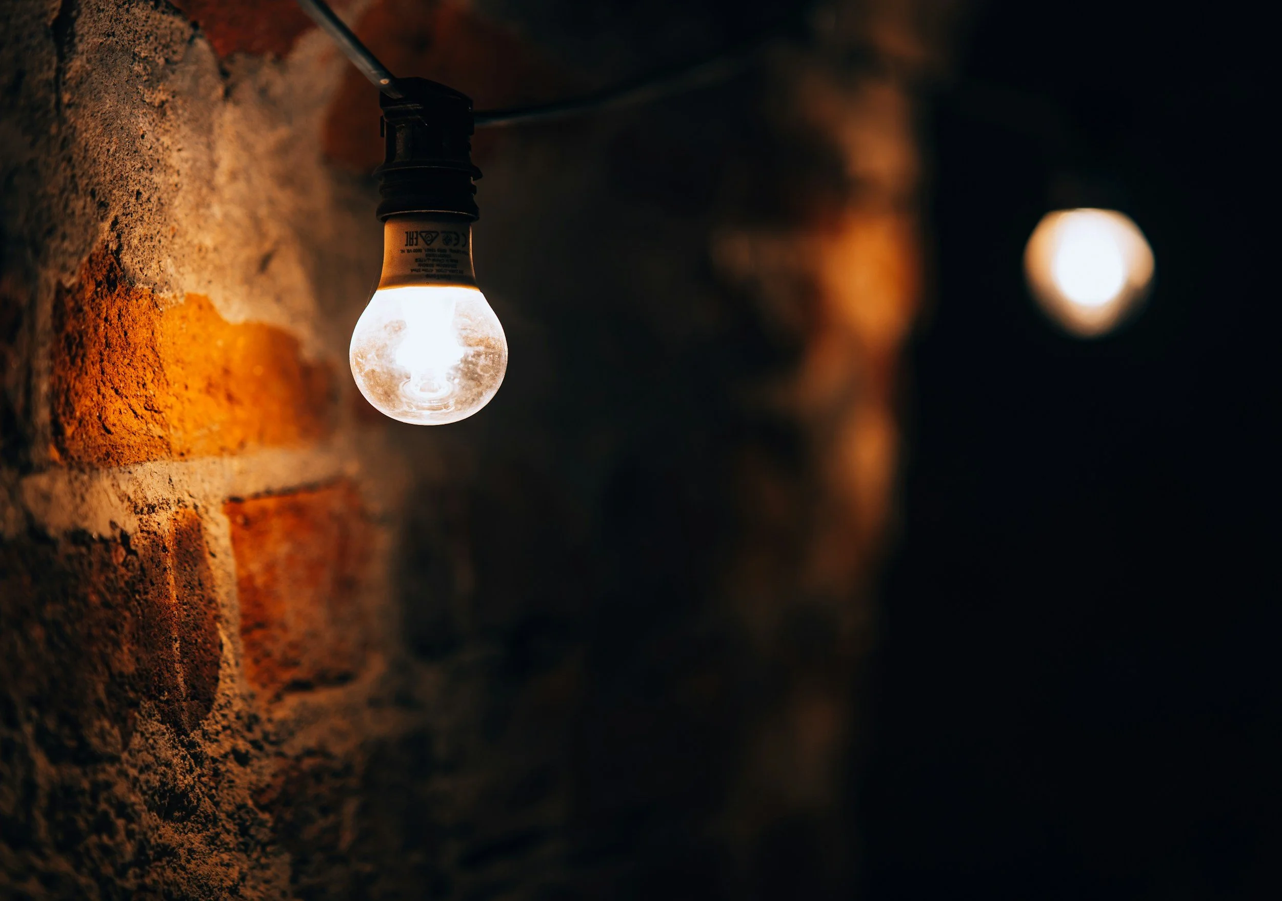 A lit lightbulb hanging on a brick wall, with a second blurred lightbulb in the background in a dark setting.