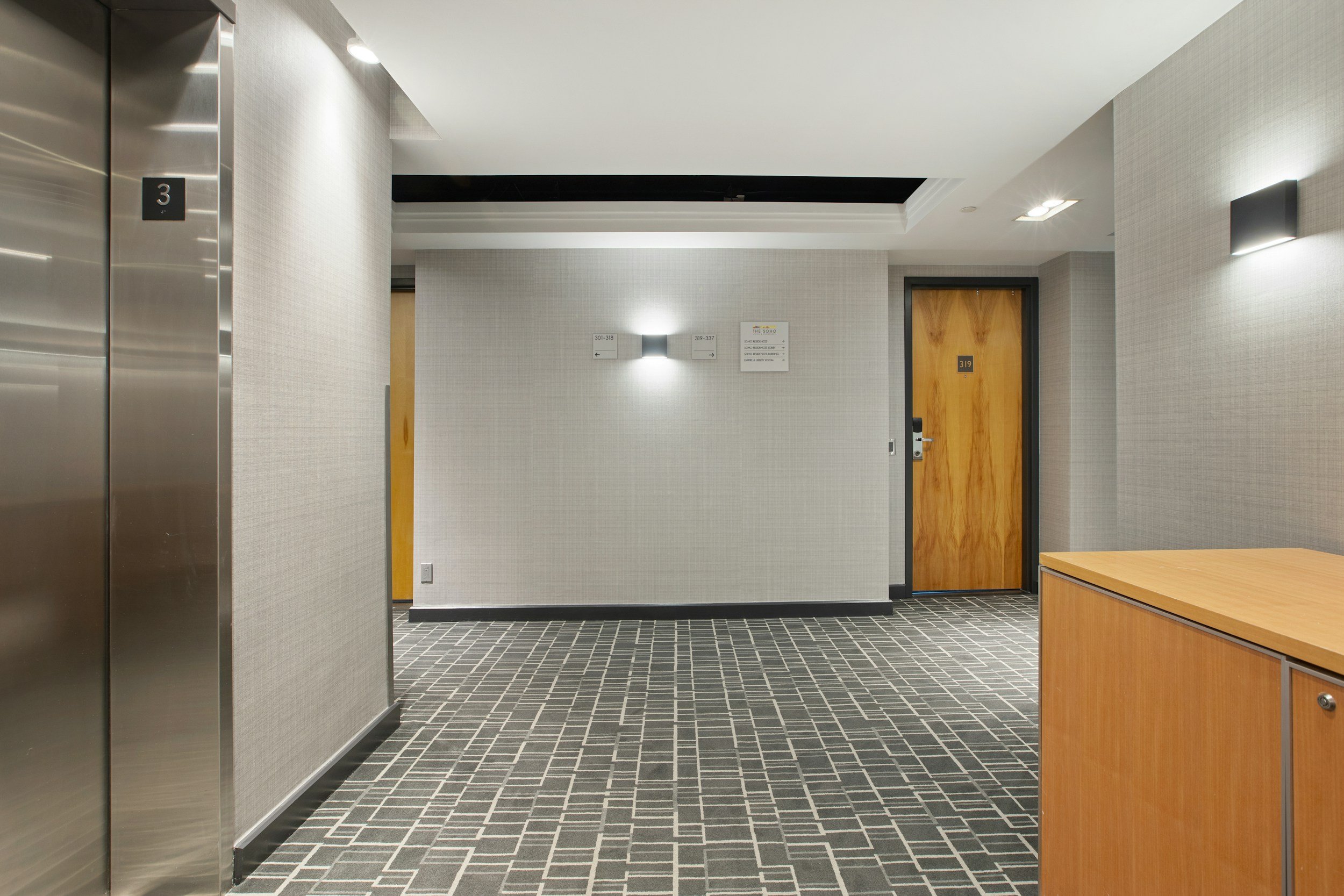 Hotel corridor with elevator and wooden doors, gray walls, patterned carpet, and wall lighting.