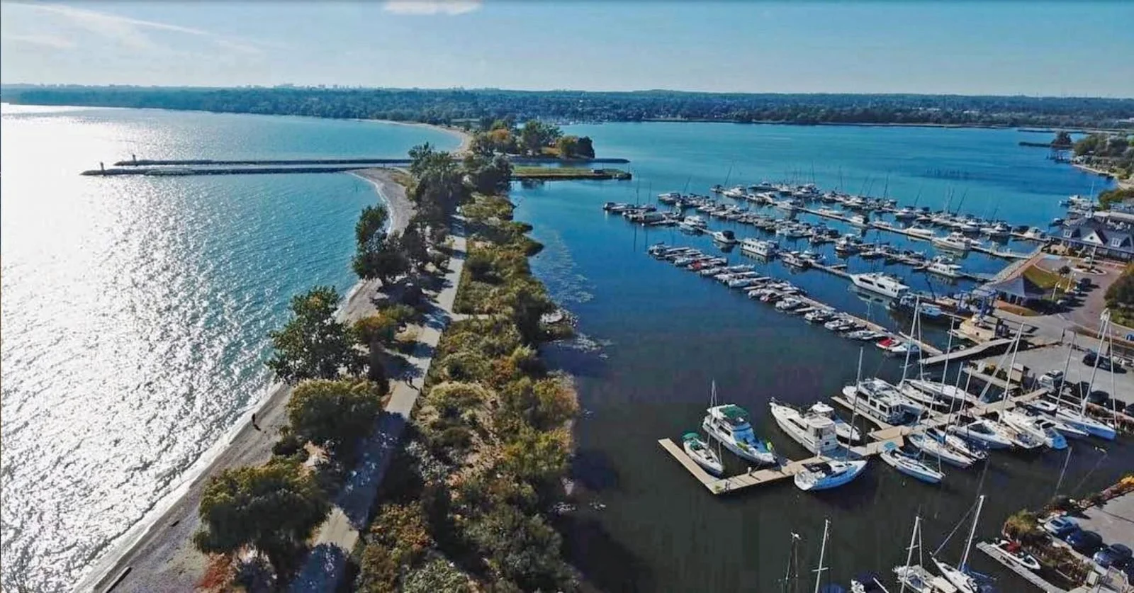 Aerial view of a marina with boats docked, a walking path lined with trees, and a large body of water reflecting sunlight, with land and a shoreline in the background.