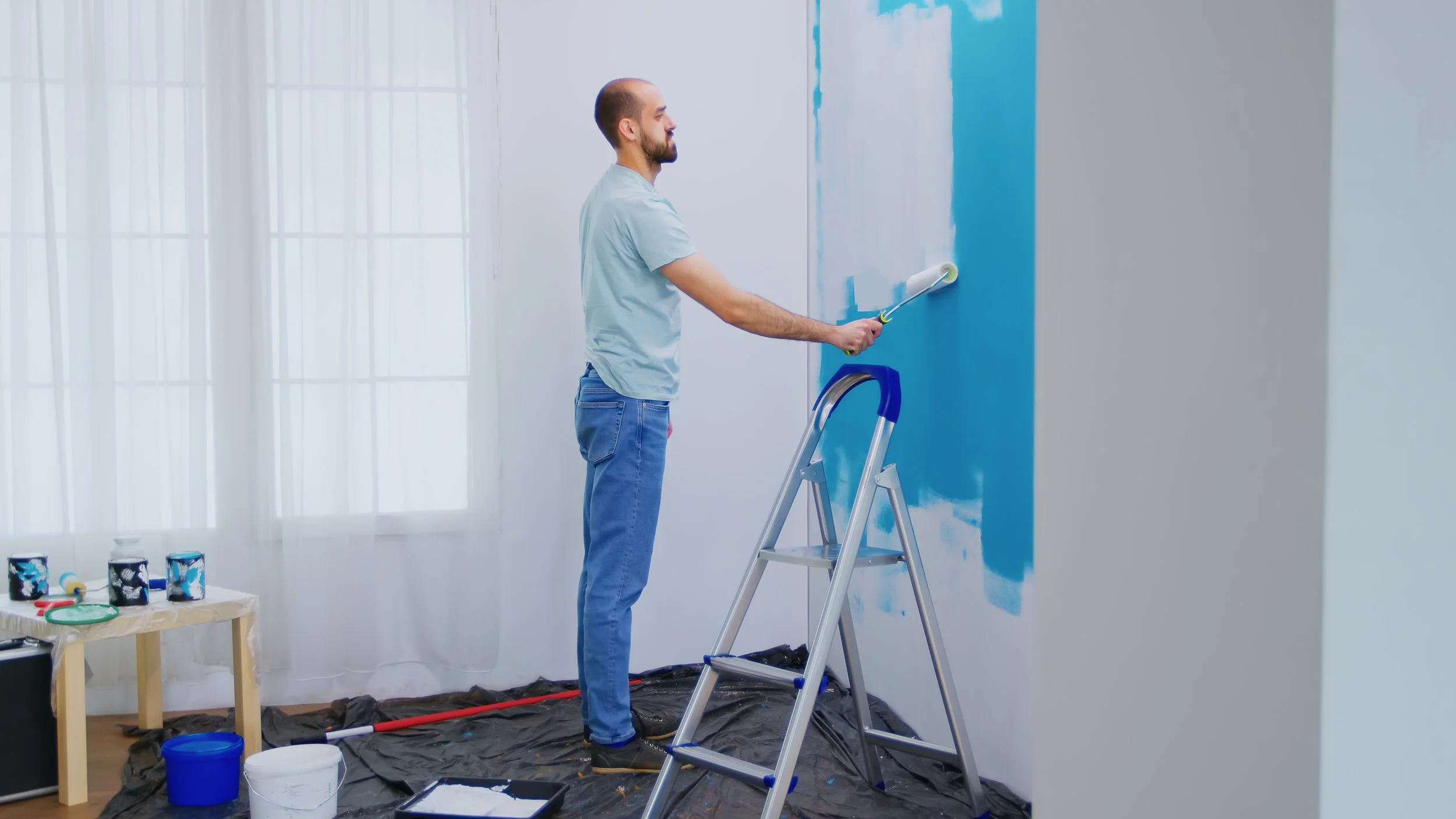 A man standing on a step stool painting a wall blue with a roller brush in a well-lit room.