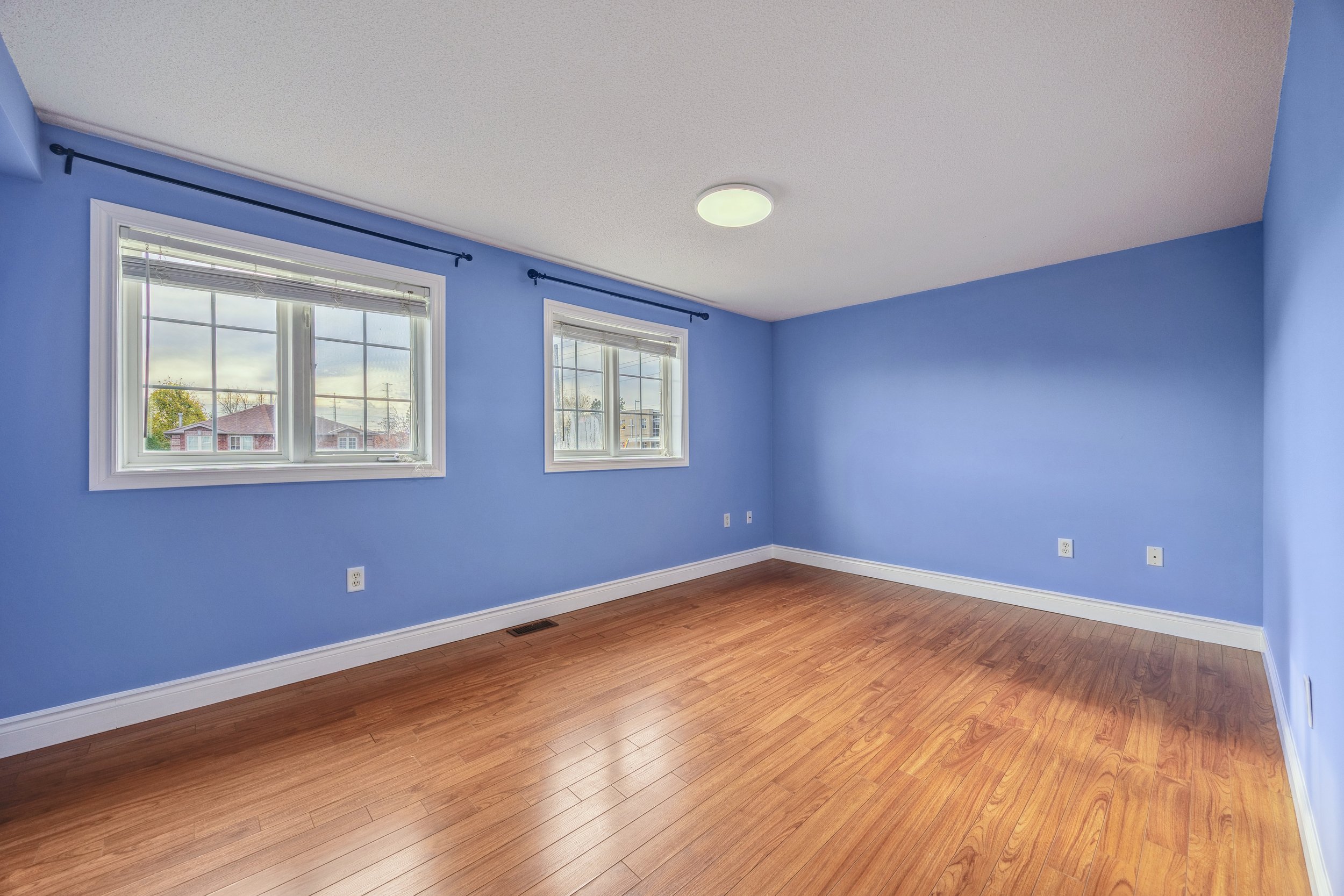 Empty room with light blue walls, hardwood floors, and two windows with white frames and blinds.