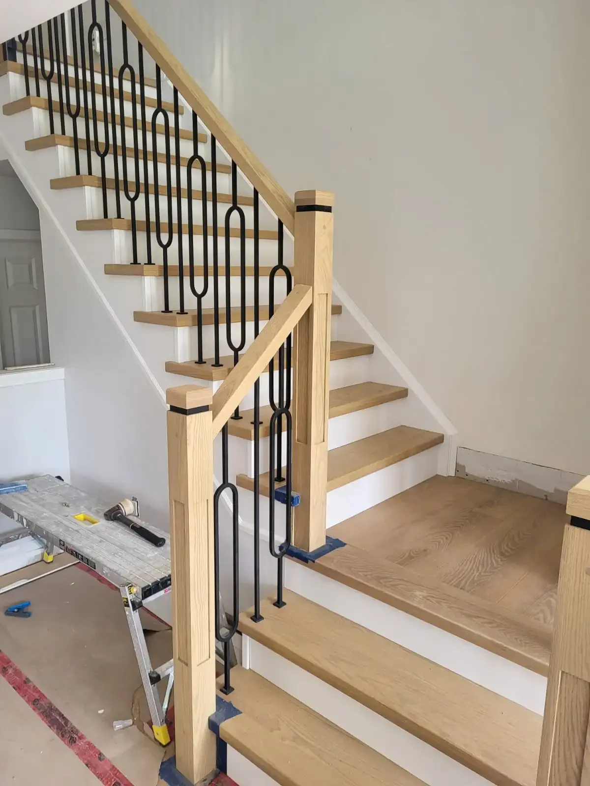 Wooden staircase with decorative black metal balusters and a newel post, under construction, with tools on the floor nearby.