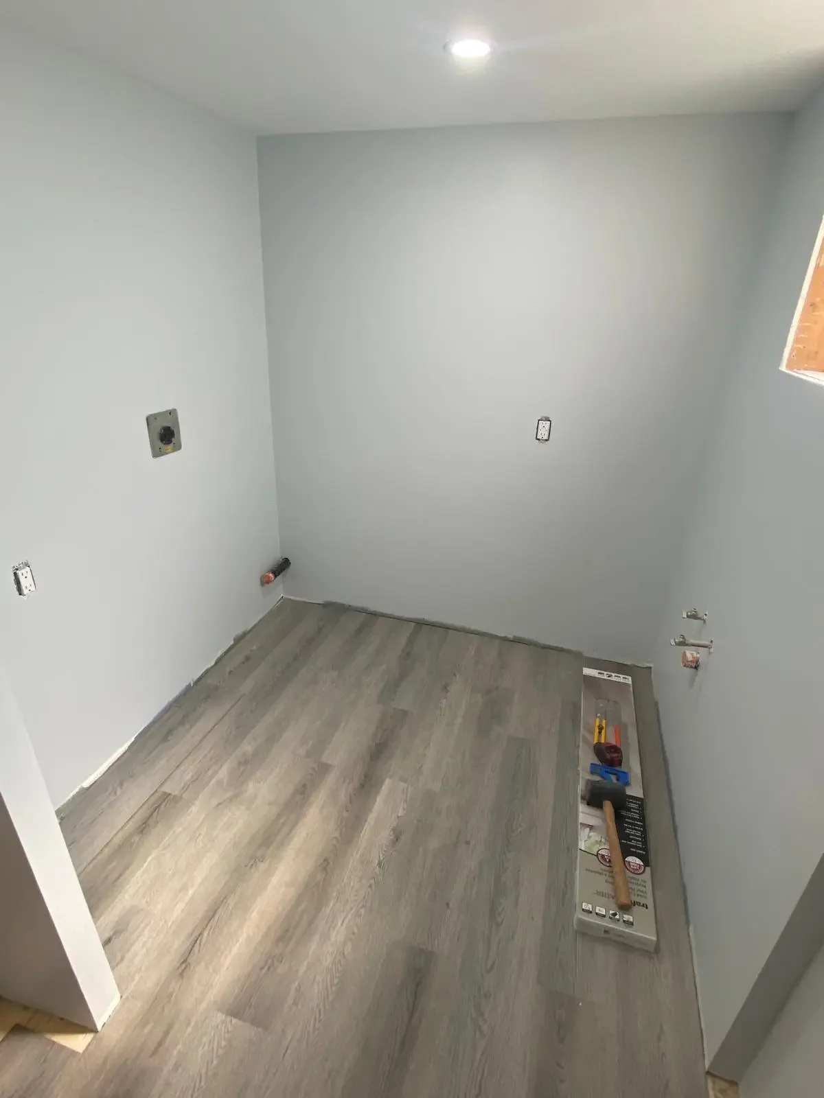 Empty room under construction with gray walls, a small window, and a new wood-look floor. Tools and drywall boxes are on the floor, with electrical outlets and plumbing fixtures visible.