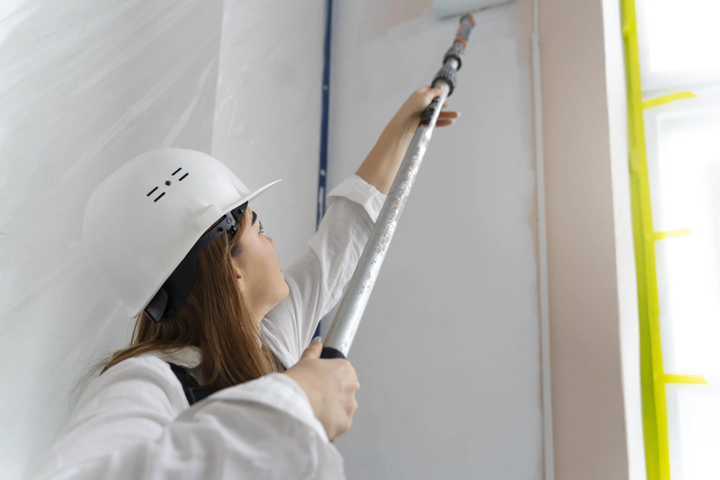 A woman wearing a white hard hat and white shirt using a level tool on a wall during construction or renovation.