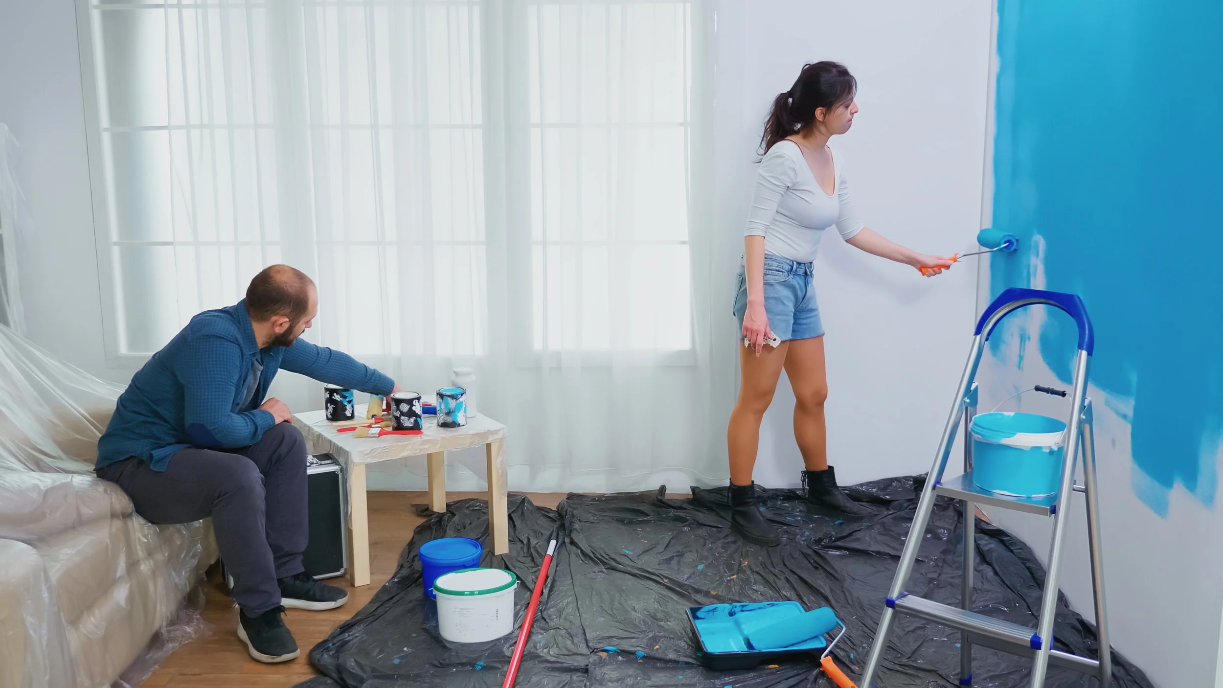 A woman is painting a wall blue with a paint roller, while a man sitting on a sofa reaches for a paintbrush and some supplies on a table nearby. The room is covered with black plastic sheeting to protect the floor, and various paint supplies are scattered around.