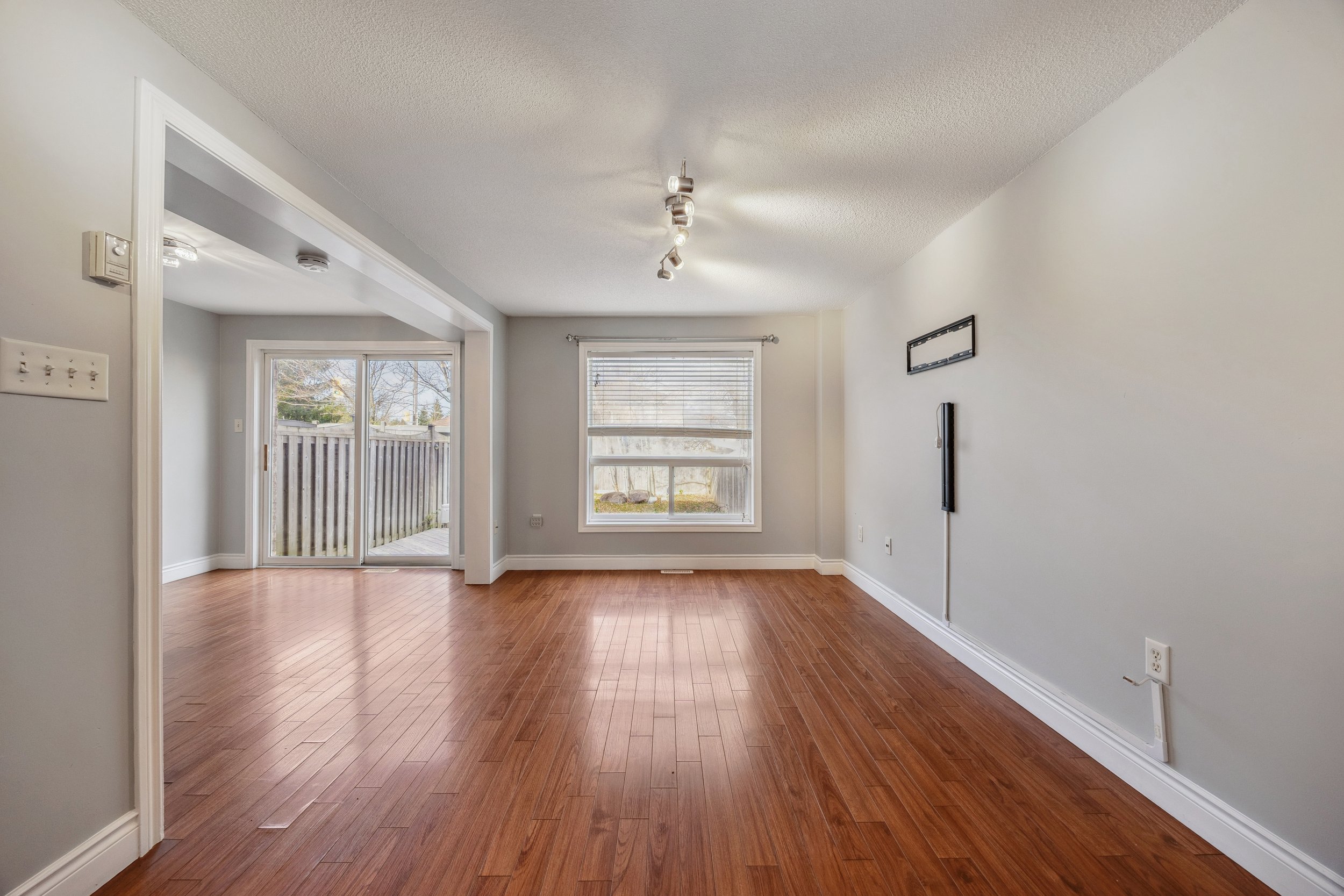 Empty living room with hardwood floors, large window, sliding glass door leading outside, and neutral-colored walls.