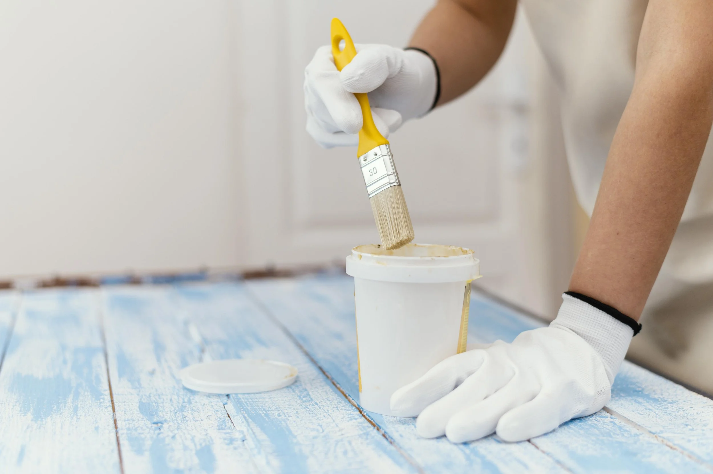 Person using a paintbrush to apply white paint or coating to a container on a wooden surface.