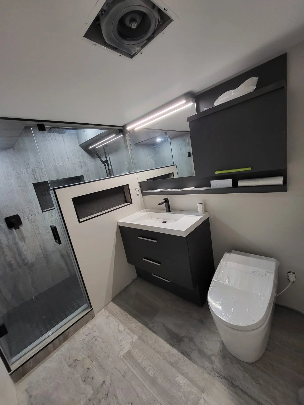 Modern bathroom with a walk-in shower, a small sink vanity with a black cabinet, a toilet with a plastic cover, a black wall-mounted cabinet, and a mirror with integrated lighting.