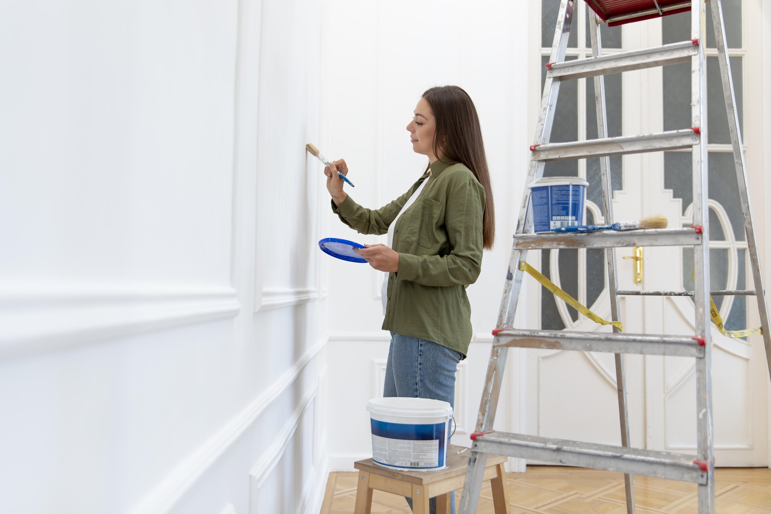 Woman painting white wall with a paintbrush while standing on the floor next to a stepladder