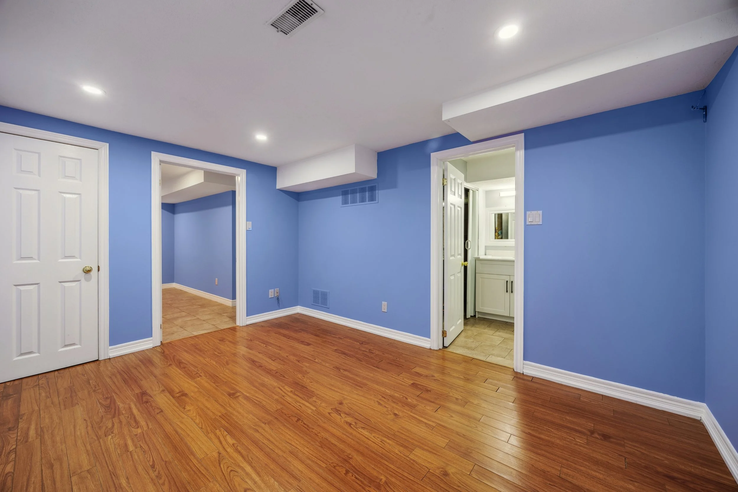 Empty living room with blue walls, hardwood flooring, and two doorways leading to other rooms. one doorway shows a bathroom with a white vanity, and the other shows a trim with beige tile flooring.