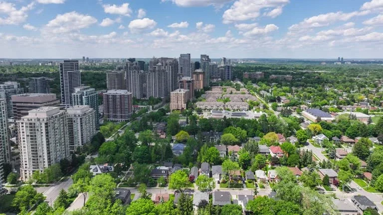 Aerial view of a city with tall modern skyscrapers and residential houses surrounded by trees on a partly cloudy day.