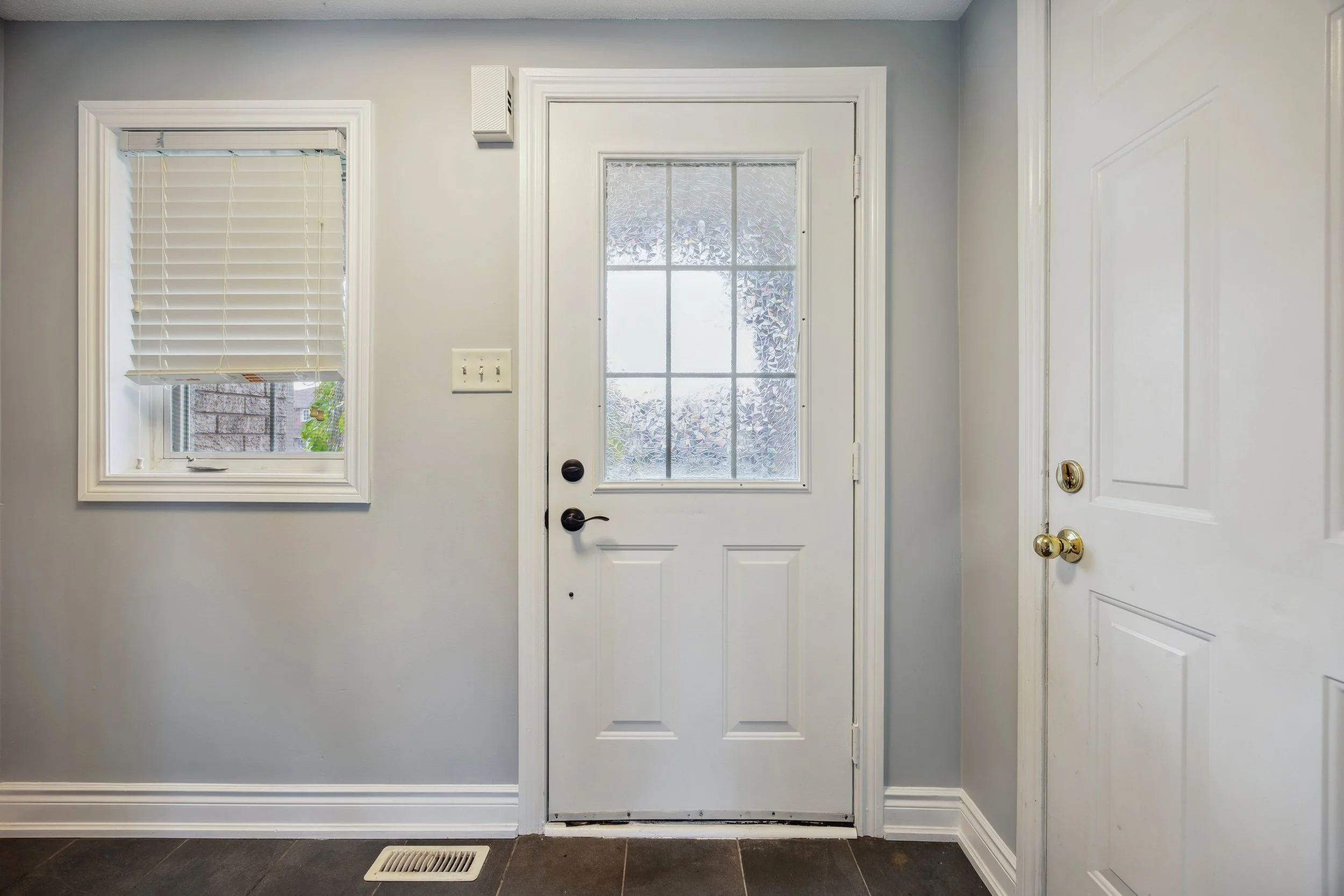 Interior view of a white door with a window, next to a window with blinds, on a gray wall with a vent and light switchplate.