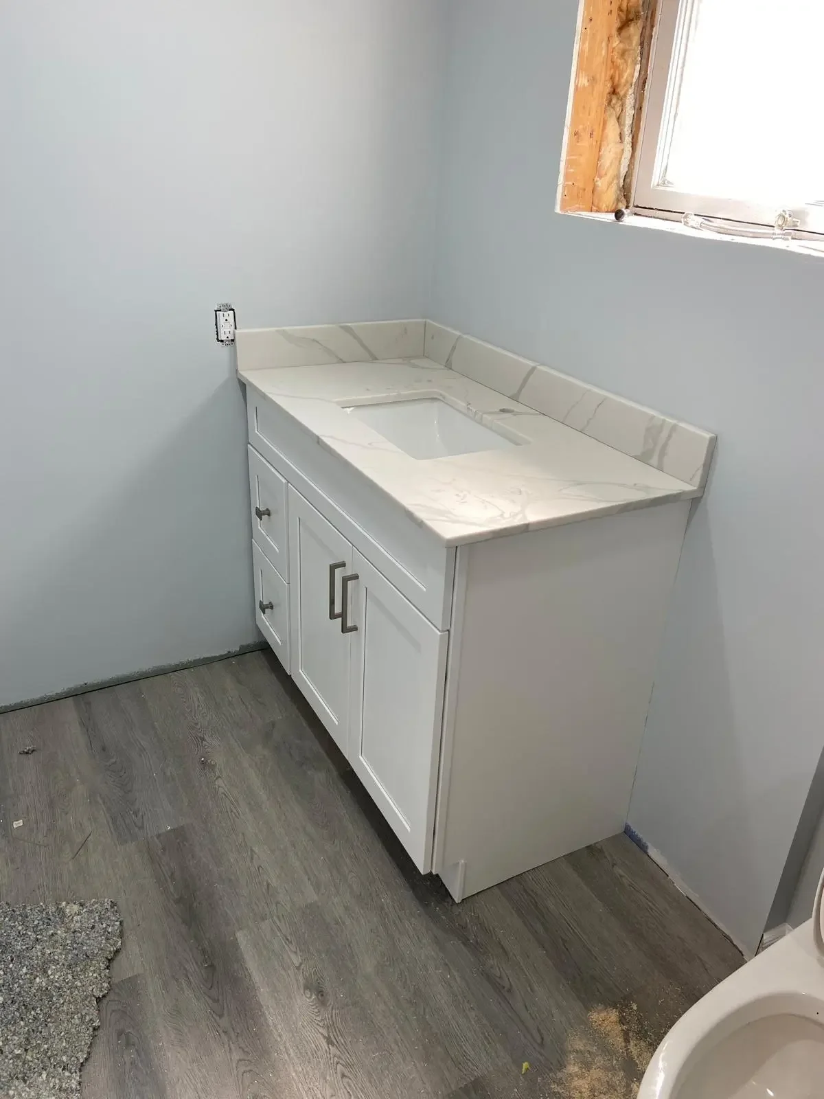 Bathroom vanity with sink and marble countertop, partially installed, with a window above and a gray wood-like floor.