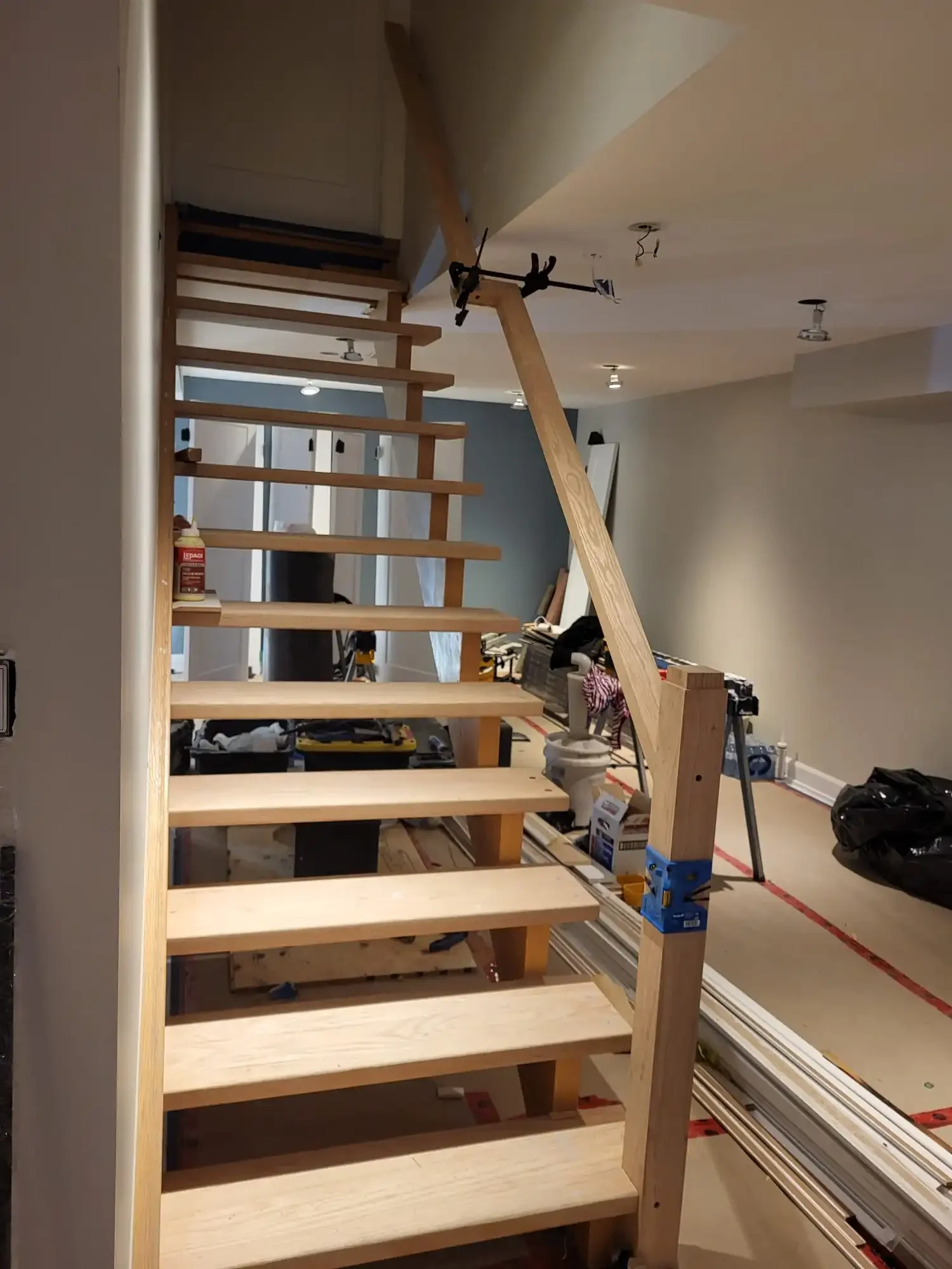 Interior view of a home under construction, showing a wooden staircase with open risers, protective blue tape on some parts, and construction tools scattered on the floor.