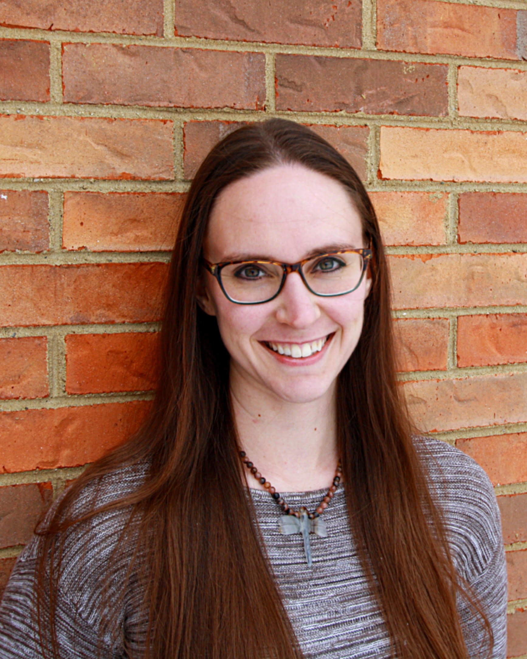 A woman with glasses and long brown hair smiling against a brick wall background.