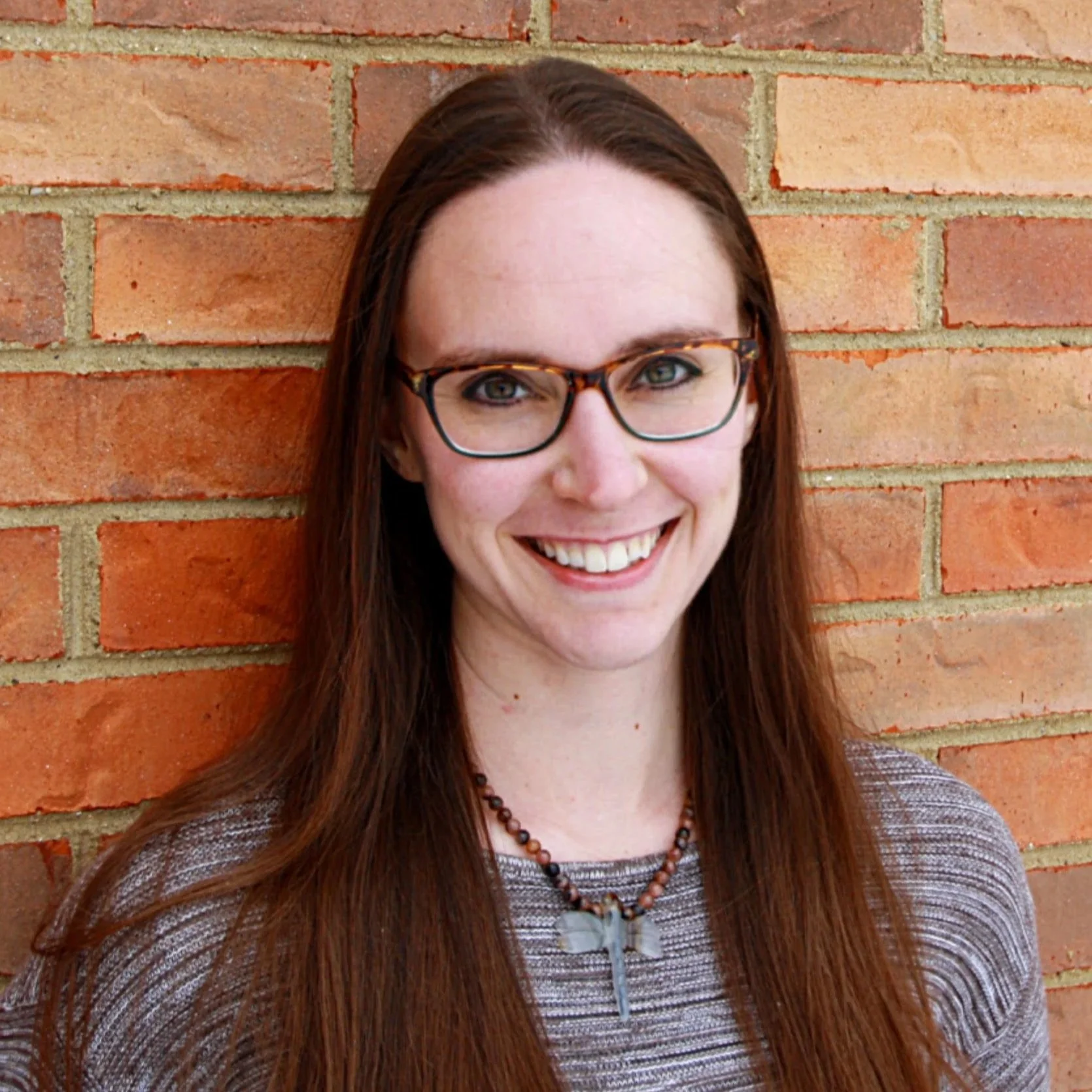 A woman with long brown hair, glasses, a dragonfly necklace, and a striped top is smiling in front of a brick wall.