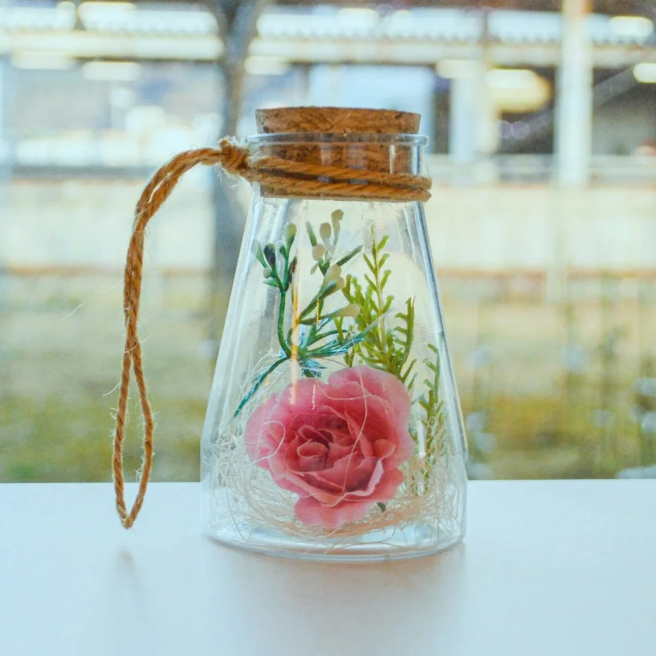 Decorative glass bottle with a pink flower and green foliage inside, sealed with a cork and tied with twine, positioned on a white surface with blurred background.