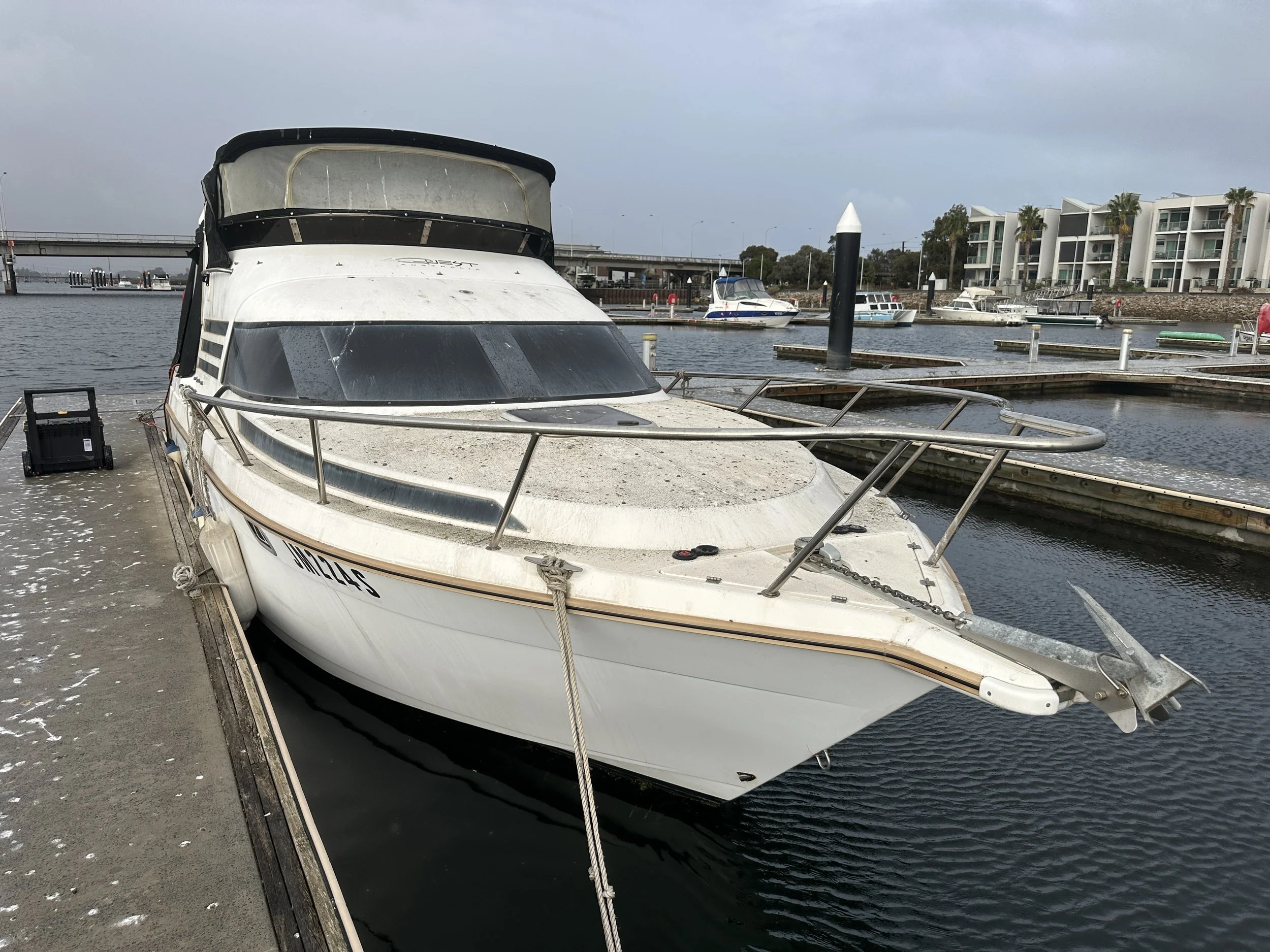 A white motor yacht docked at a marina with other boats and modern apartment buildings in the background.