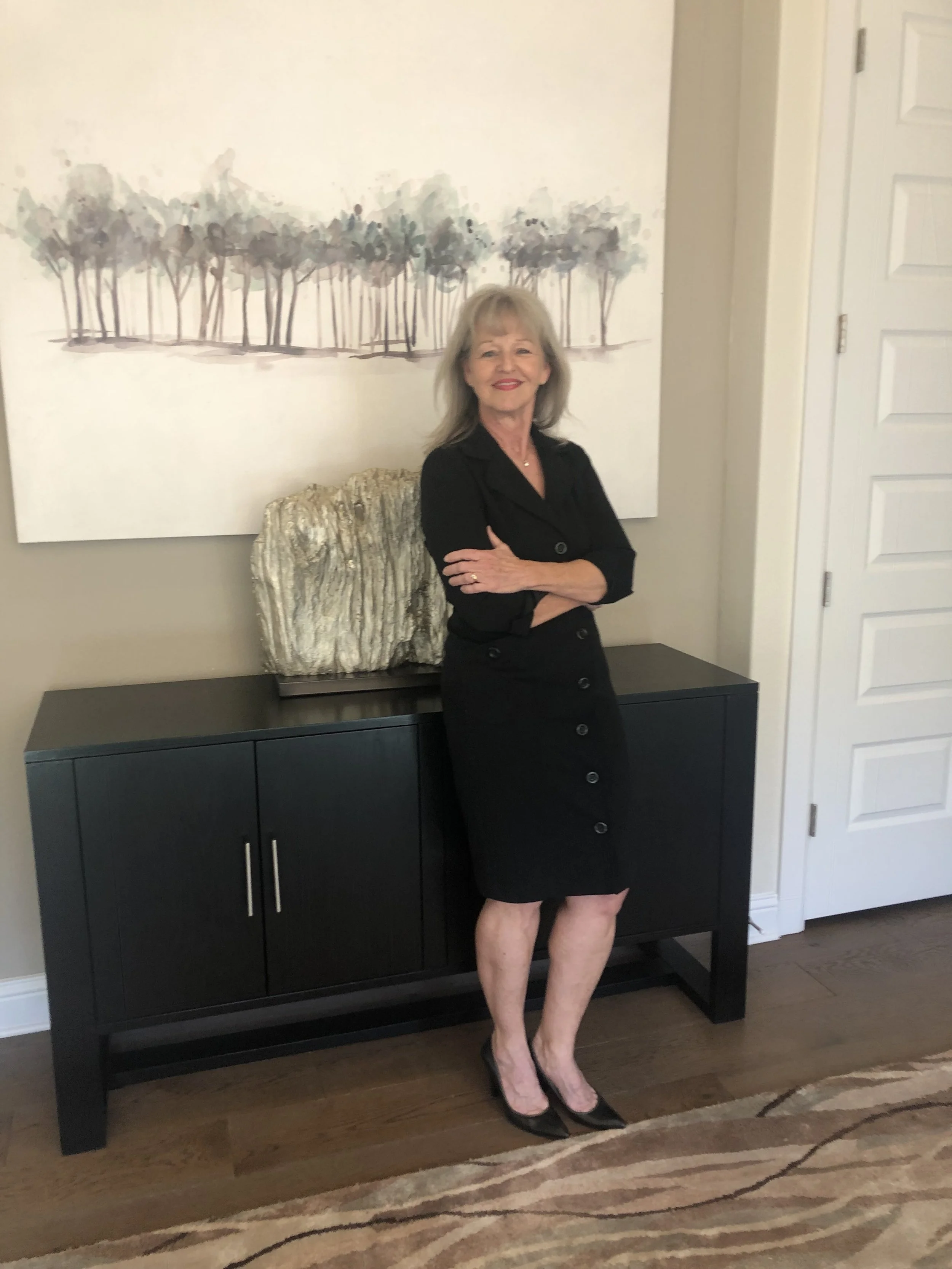 A woman with shoulder-length gray hair wearing a black dress and black heels standing in front of a black cabinet with her arms crossed. There is a large painting of trees on the wall behind her.