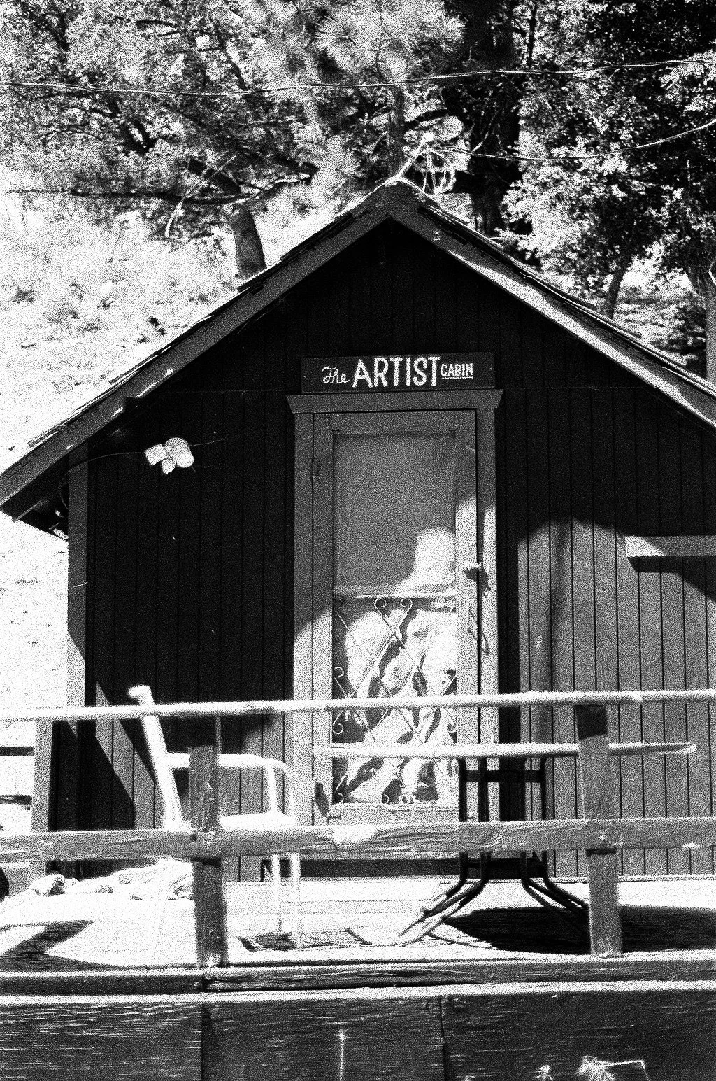 Black and white photo of a small wooden cabin labeled "The Artist Cabin" with a sign above the door, a porch with railings, a butterfly decoration on the wall, and trees in the background.