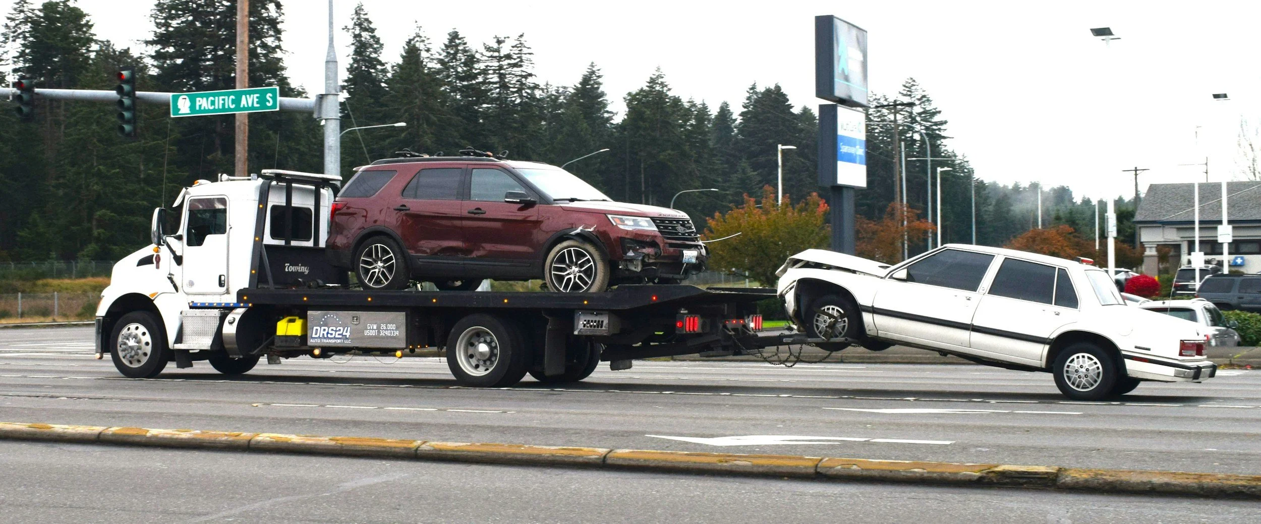 A white tow truck on a city street carrying a damaged maroon SUV and an upside-down white station wagon, with traffic lights, street signs, and buildings in the background.
