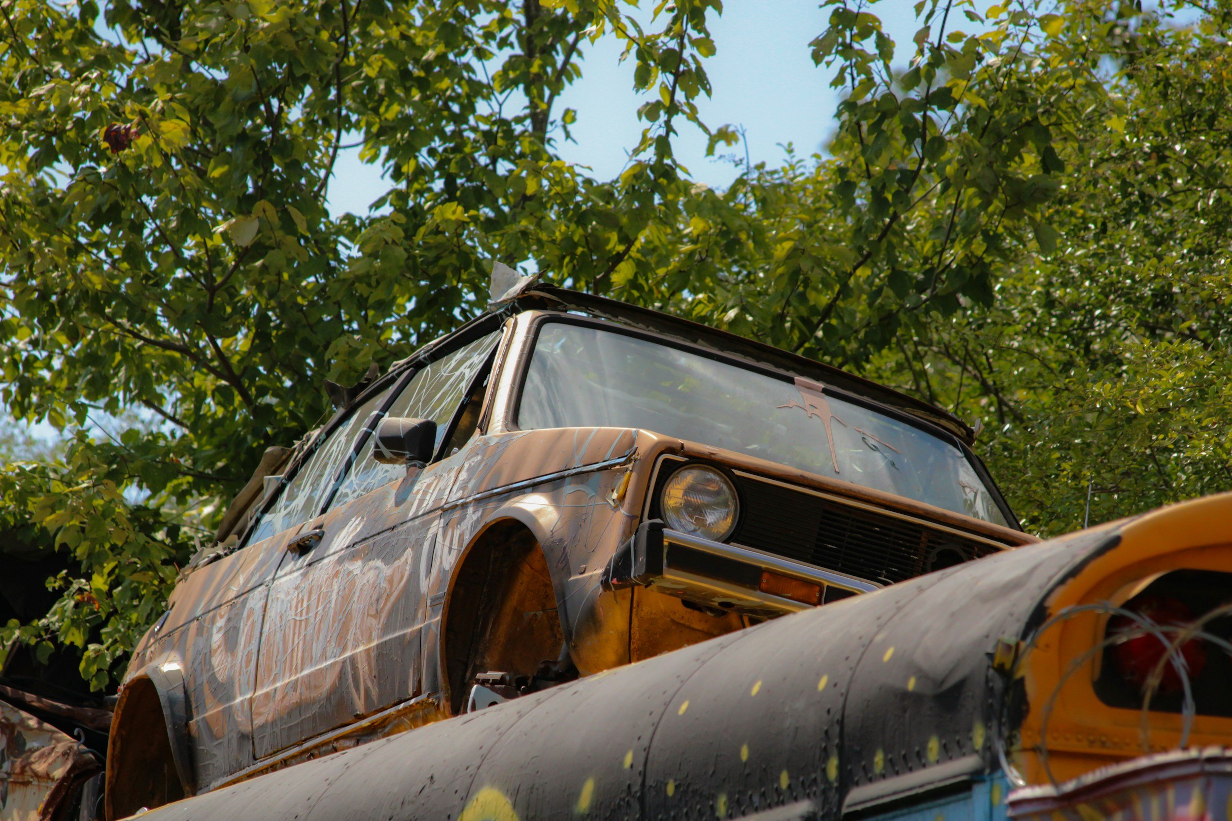 A rusty, abandoned car with a shattered windshield and dented body sitting outdoors among green trees.