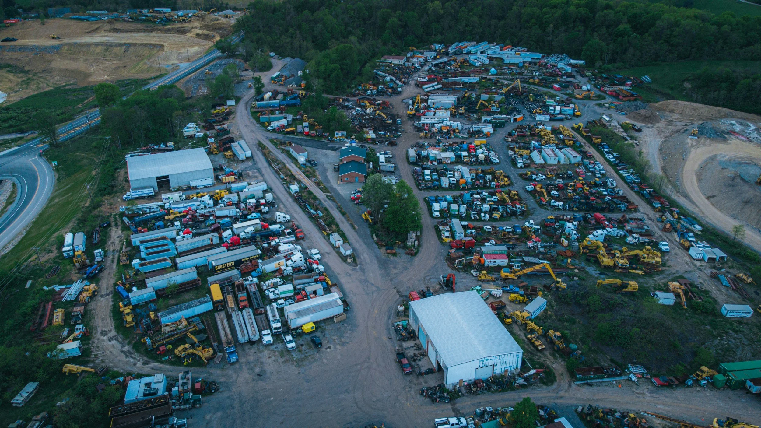An aerial view of a large junkyard with numerous trucks, construction equipment, and vehicles, surrounded by trees and construction areas.