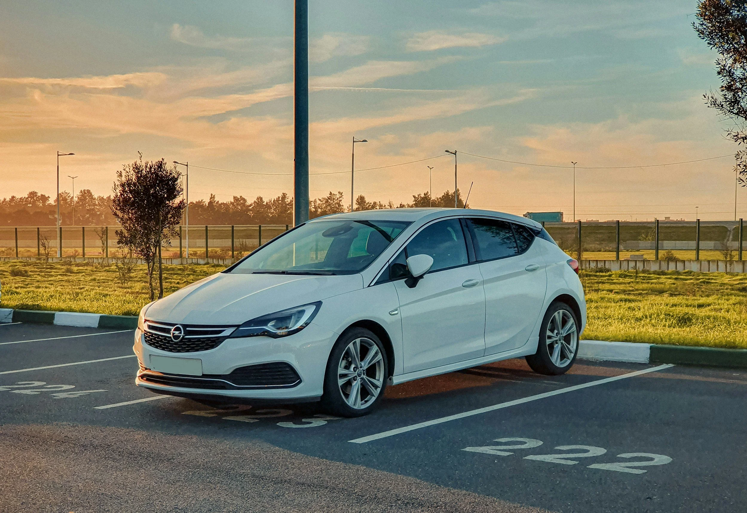 White Opel hatchback car parked in a marked parking space at sunset.
