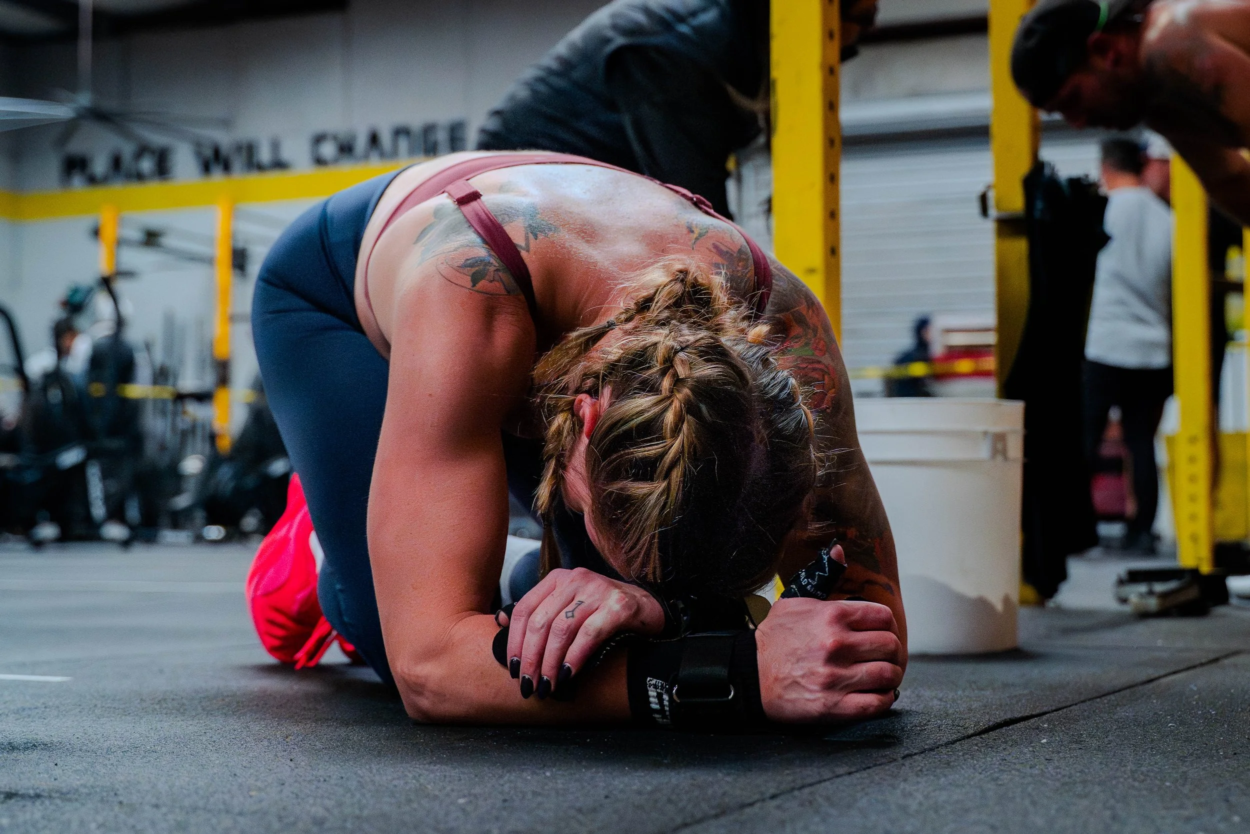 A woman with braided hair kneeling on the floor in a gym, holding her hands together in a prayer position, with other people and gym equipment visible in the background.