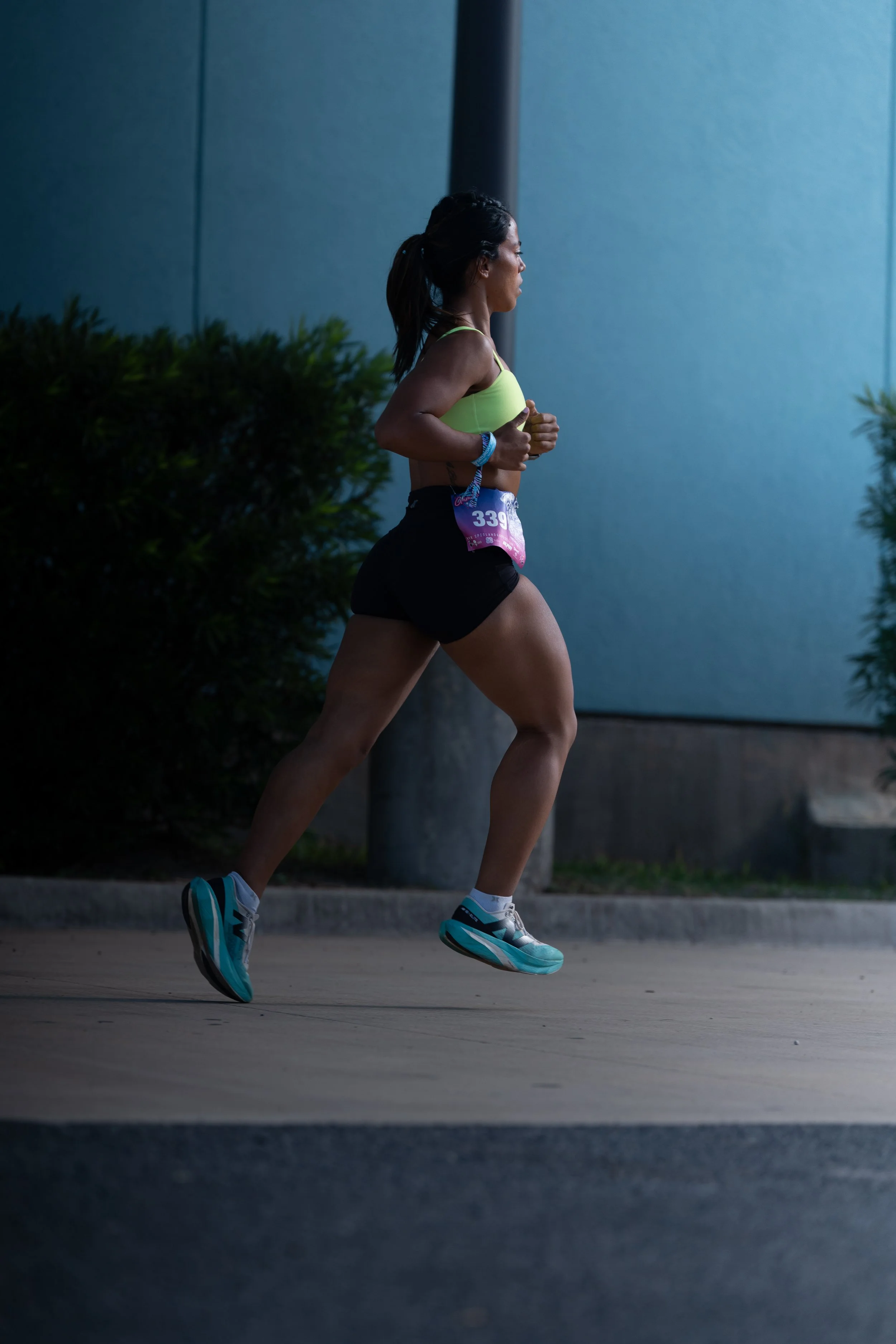 A woman running outdoors during a race, wearing a yellow sports bra, black shorts, and teal running shoes, with a race bib number 339.