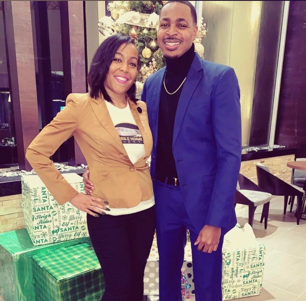 A woman and a man smiling for the camera at a Christmas-themed indoor setting, standing in front of decorated Christmas trees and wrapped presents.