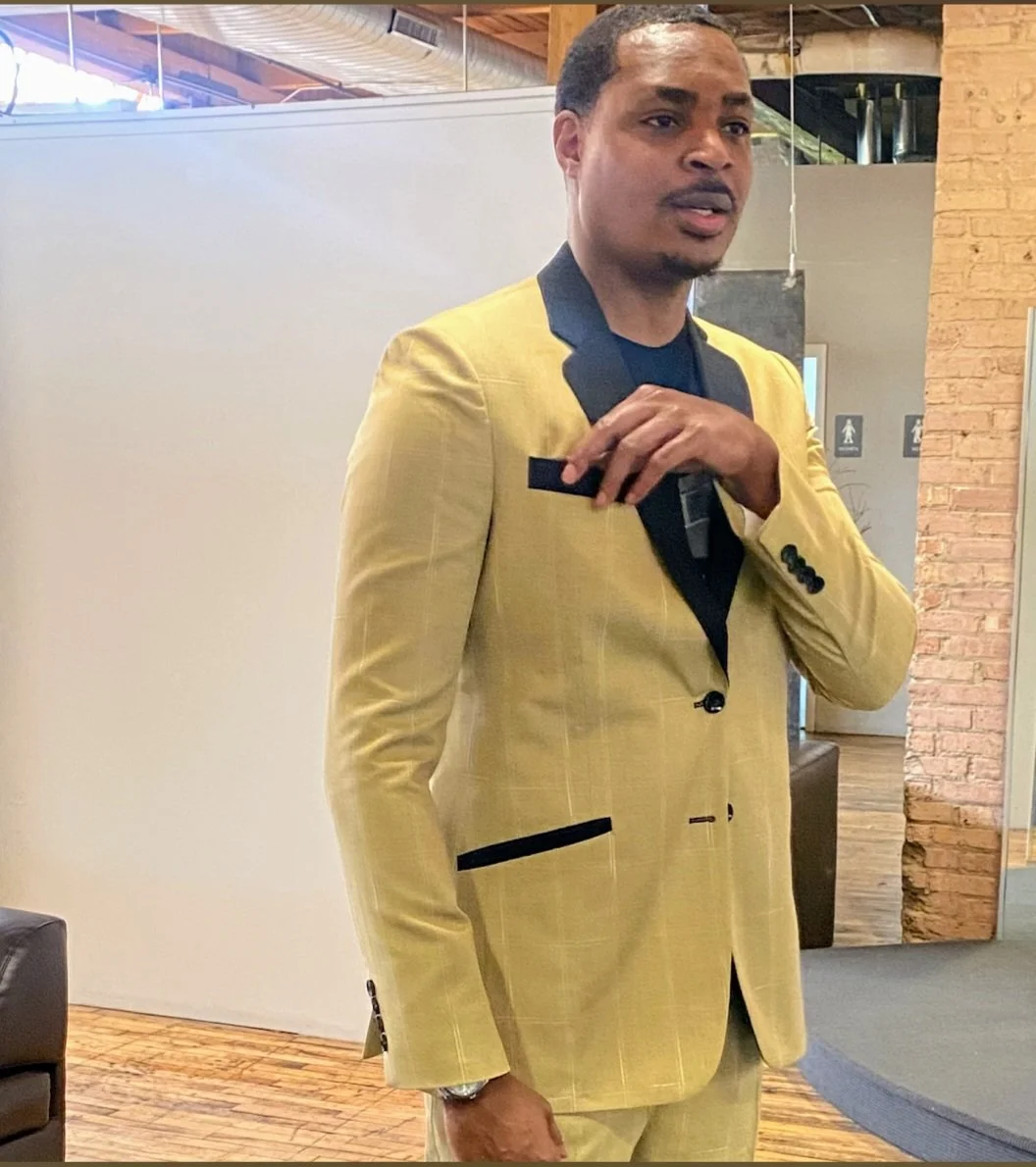 A man wearing a yellow suit with black accents is taking a mirror selfie in a modern interior space with wooden and brick elements.