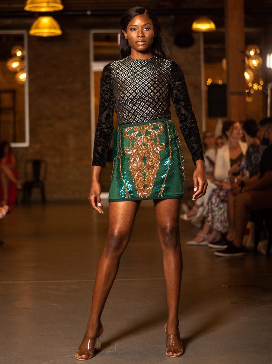 A Black woman walking down a runway during a fashion show, wearing a long-sleeved black top with a shiny geometric pattern and a green mini skirt with gold embroidery and sequins, and clear high heels. Audience members are seated on either side of the runway.