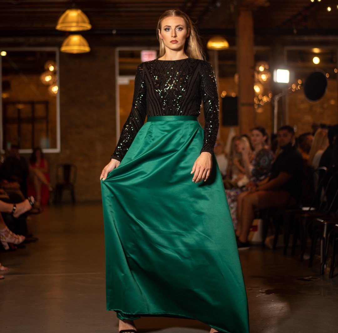 Model wearing a black sequined top and a long green satin skirt walking on runway at fashion show.