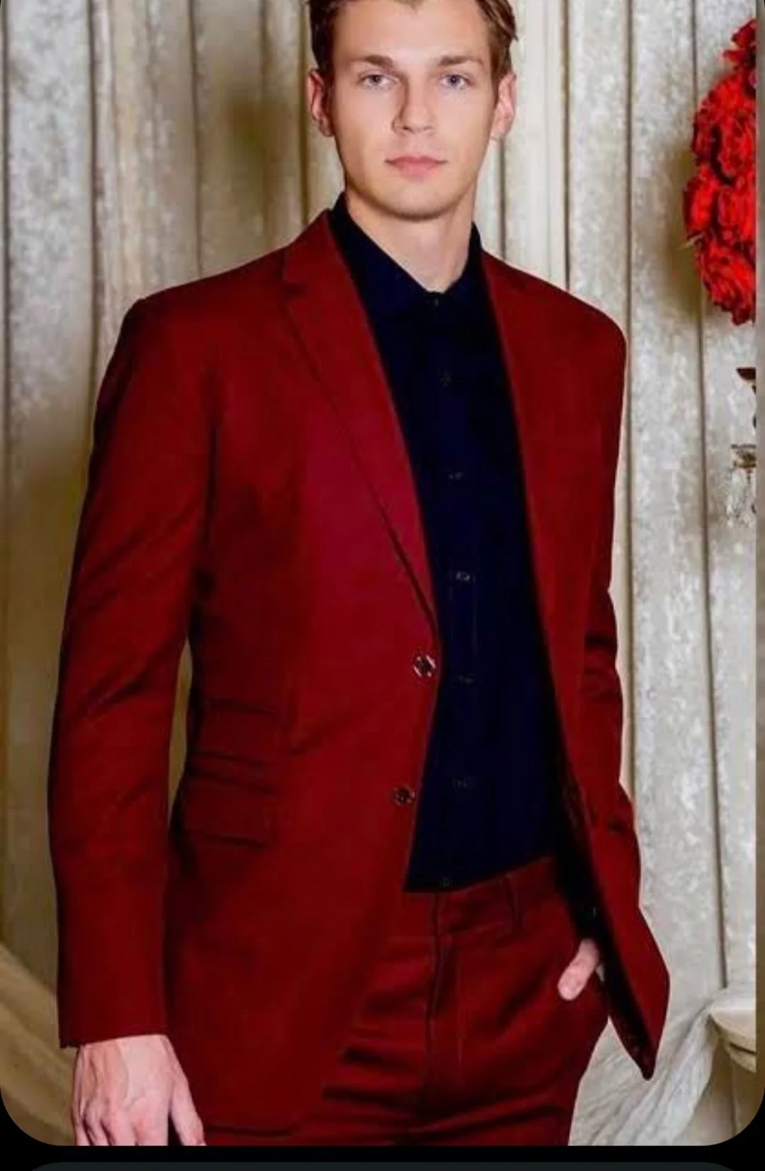 A young man in a red suit standing indoors with a serious expression, posed with one big hand in his pocket, against a beige textured background with a floral wreath to his right.