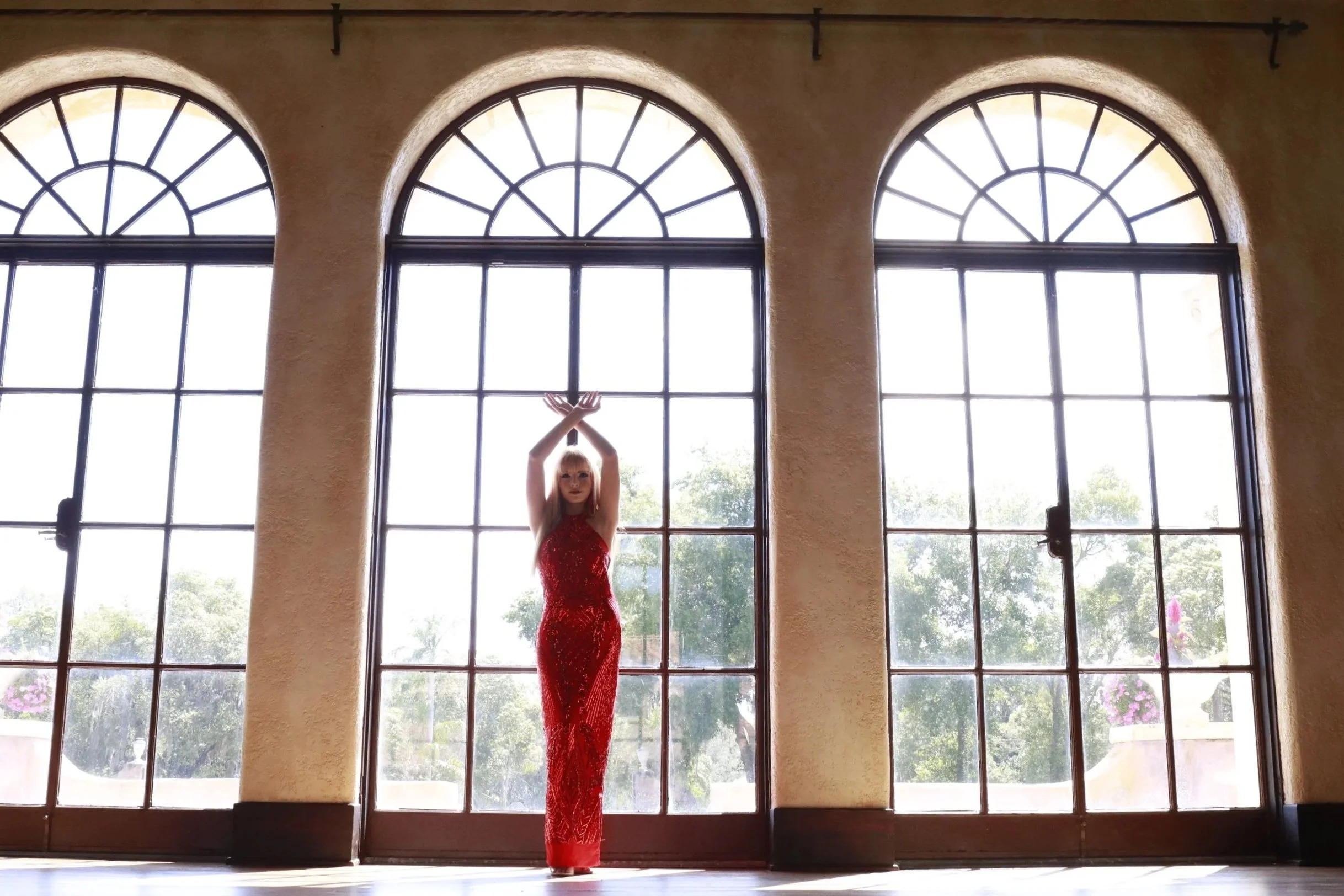 Woman in a red sequined dress standing in front of large arched windows with sunlight streaming in.