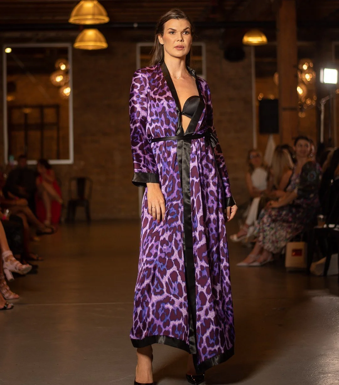 A woman wearing a purple leopard print long robe with black trim, black lingerie underneath, walking on a runway in a dimly lit venue with hanging lights and an audience.
