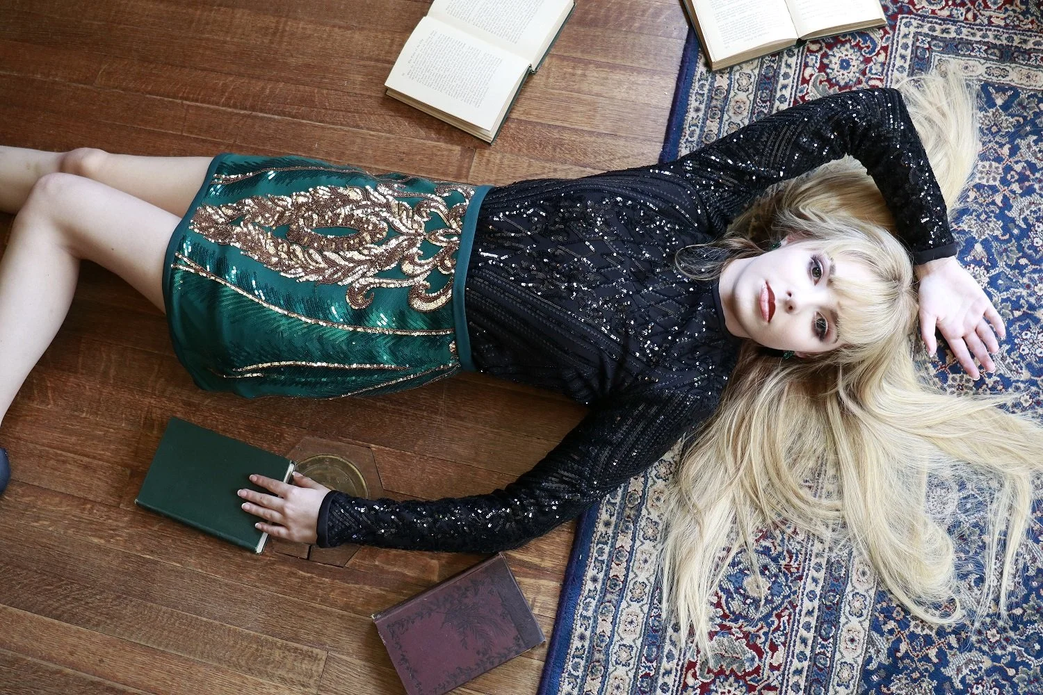 A woman with long blonde hair lying on a wooden floor, wearing a sparkling black top and a decorative green and gold skirt, surrounded by open books and a closed notebook.