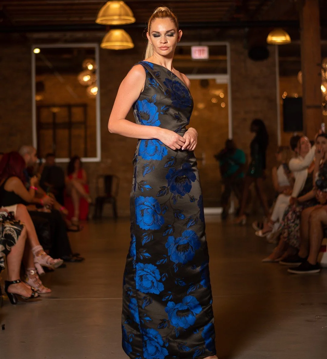 Woman in a long black dress with blue floral pattern walking on runway at fashion show.